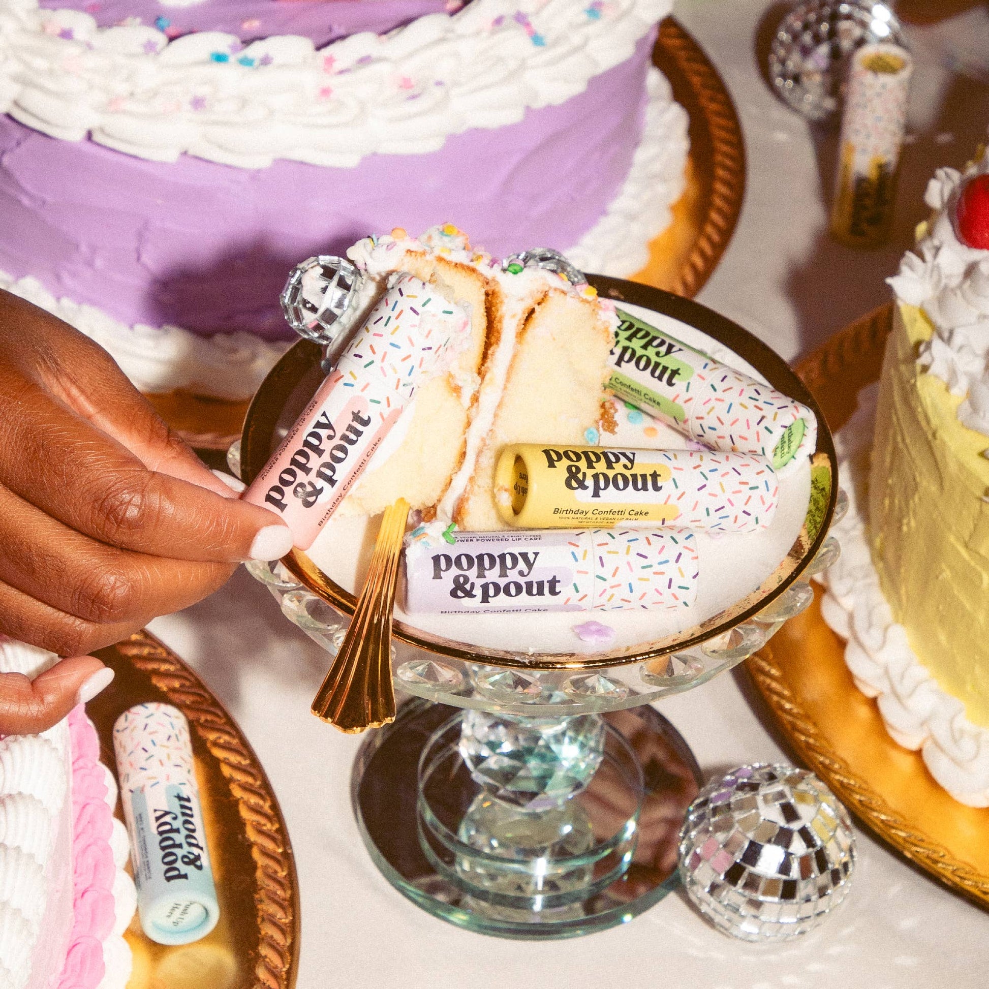 Hand holding a small cake with 'Poppy & Pout' branding, surrounded by larger cakes and decorative items.