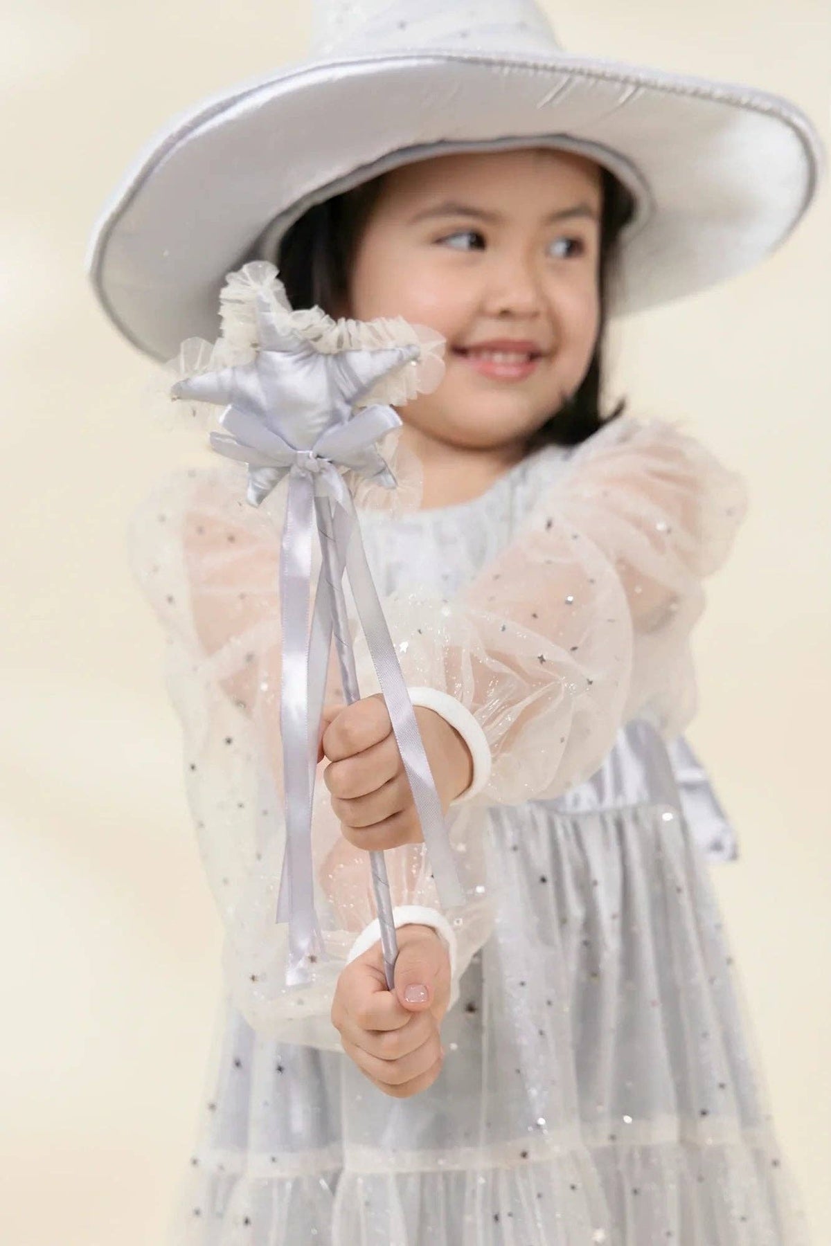 A young girl wearing a white wide-brim hat and a sparkly white tulle dress holds a matching white and silver bow-tied wand against a soft beige background.