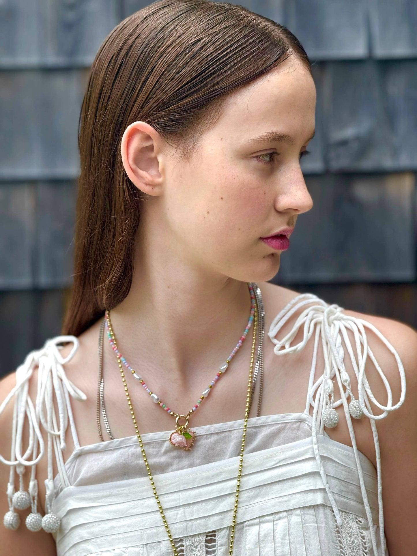 Woman wearing a white dress with fringe details and multiple necklaces against a wooden wall.