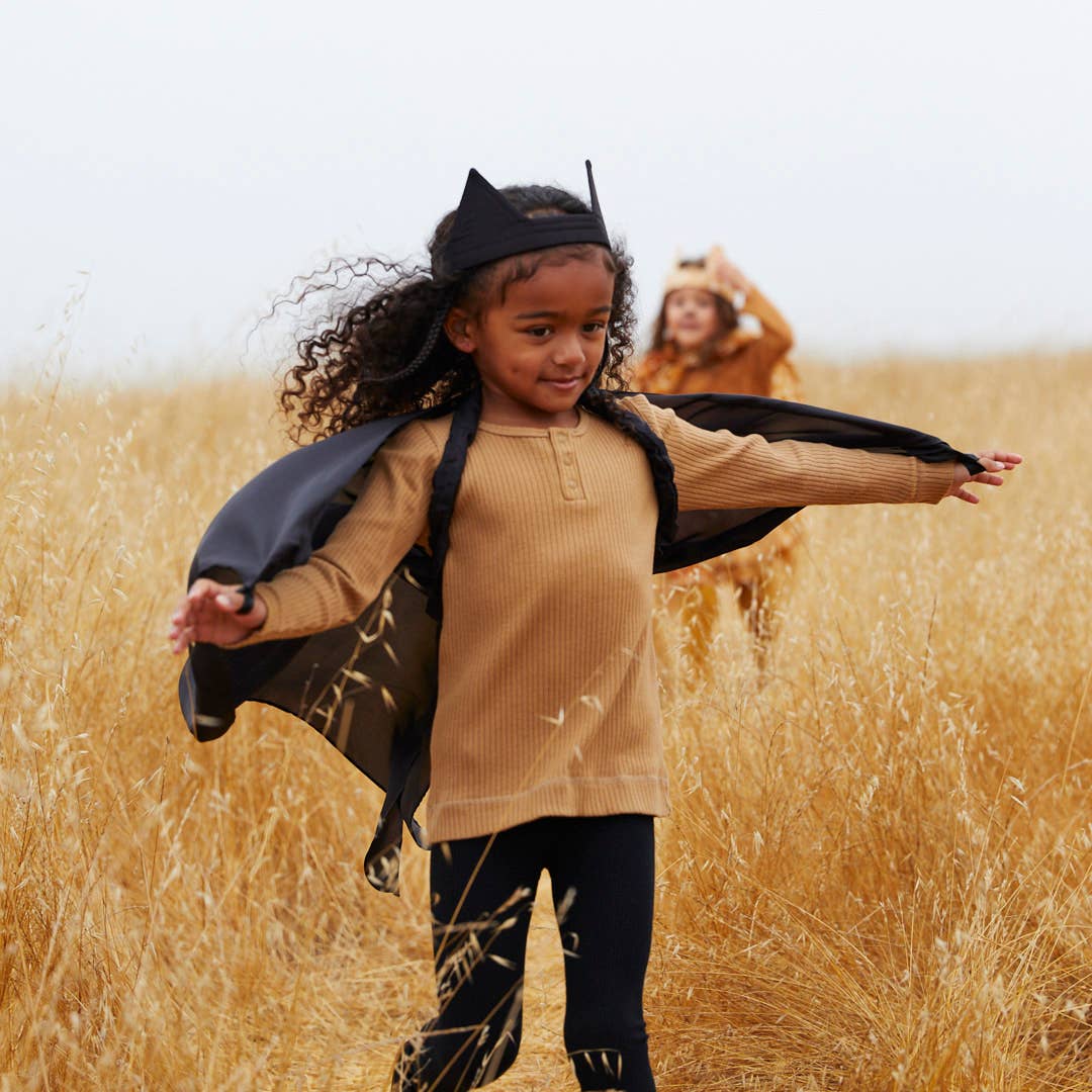 A young girl wearing a tan ribbed henley shirt, black leggings, and a black cat cape stands in a golden field, with a child in a dog costume blurred in the background.