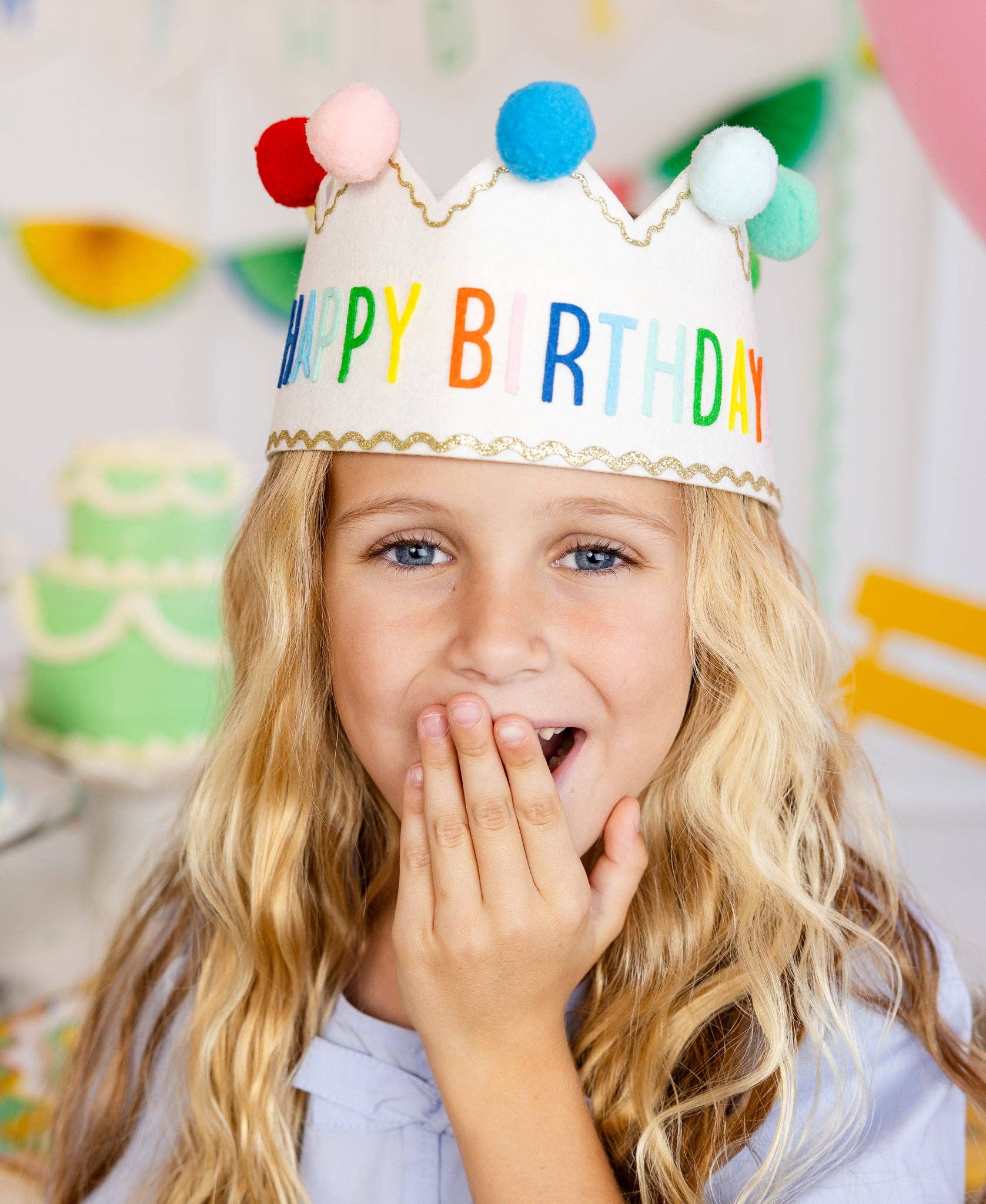 Child wearing a colorful 'Happy Birthday' crown with a blurred birthday cake and decorations in the background.