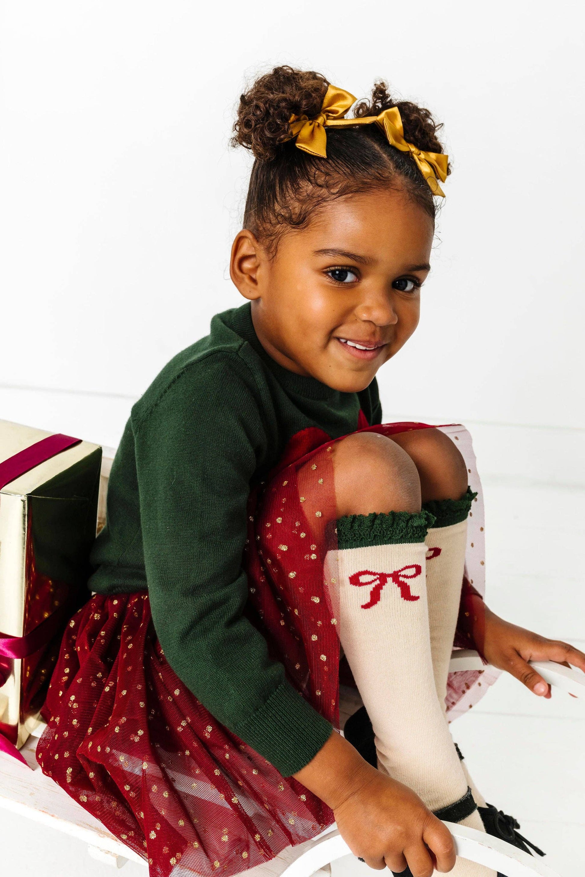 Young girl in a green sweater and red dress with white knee-high socks, sitting on a white surface.