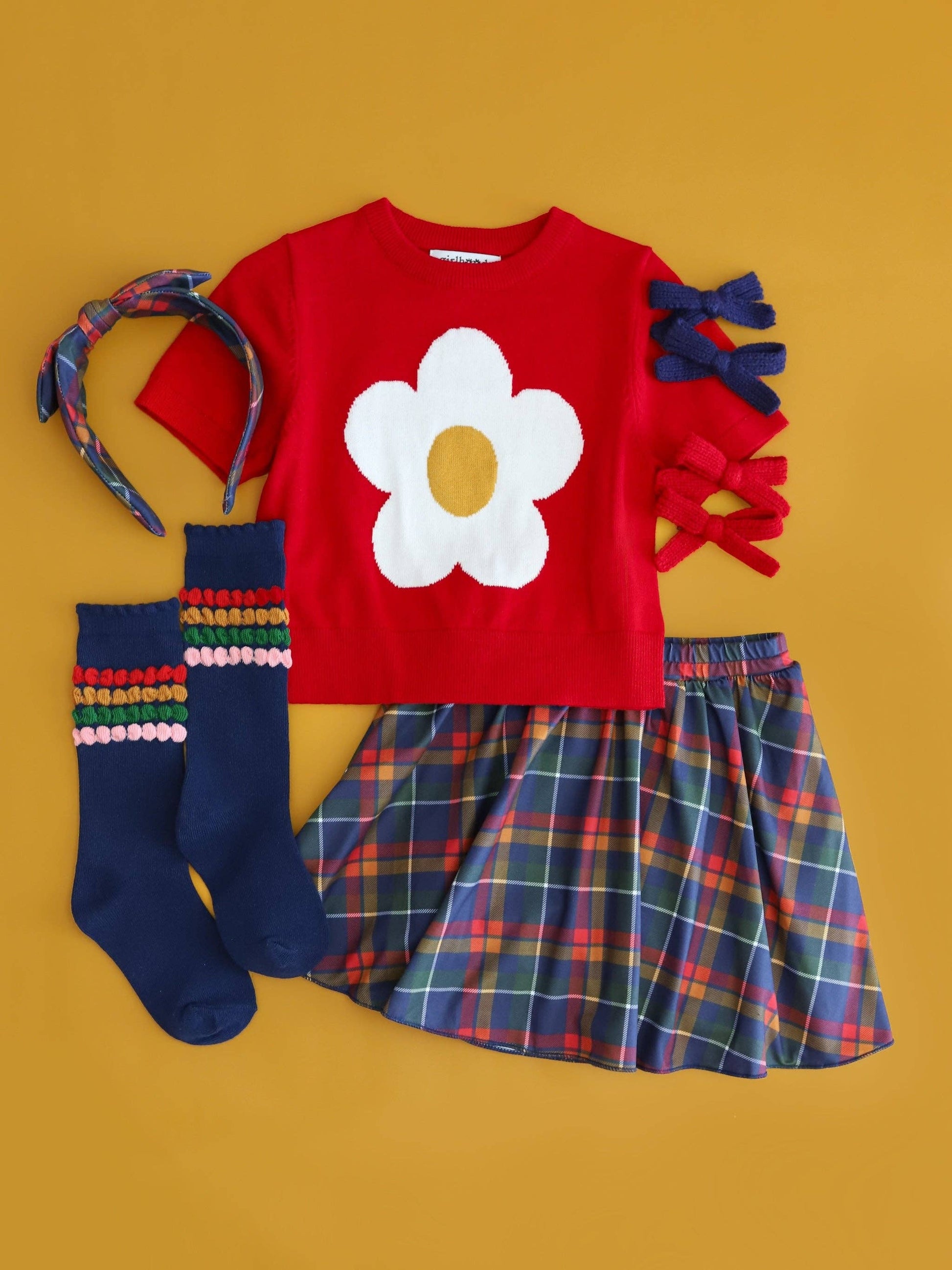 Children's outfit with red shirt with flower design, plaid skirt, colorful socks, and headband on a yellow background