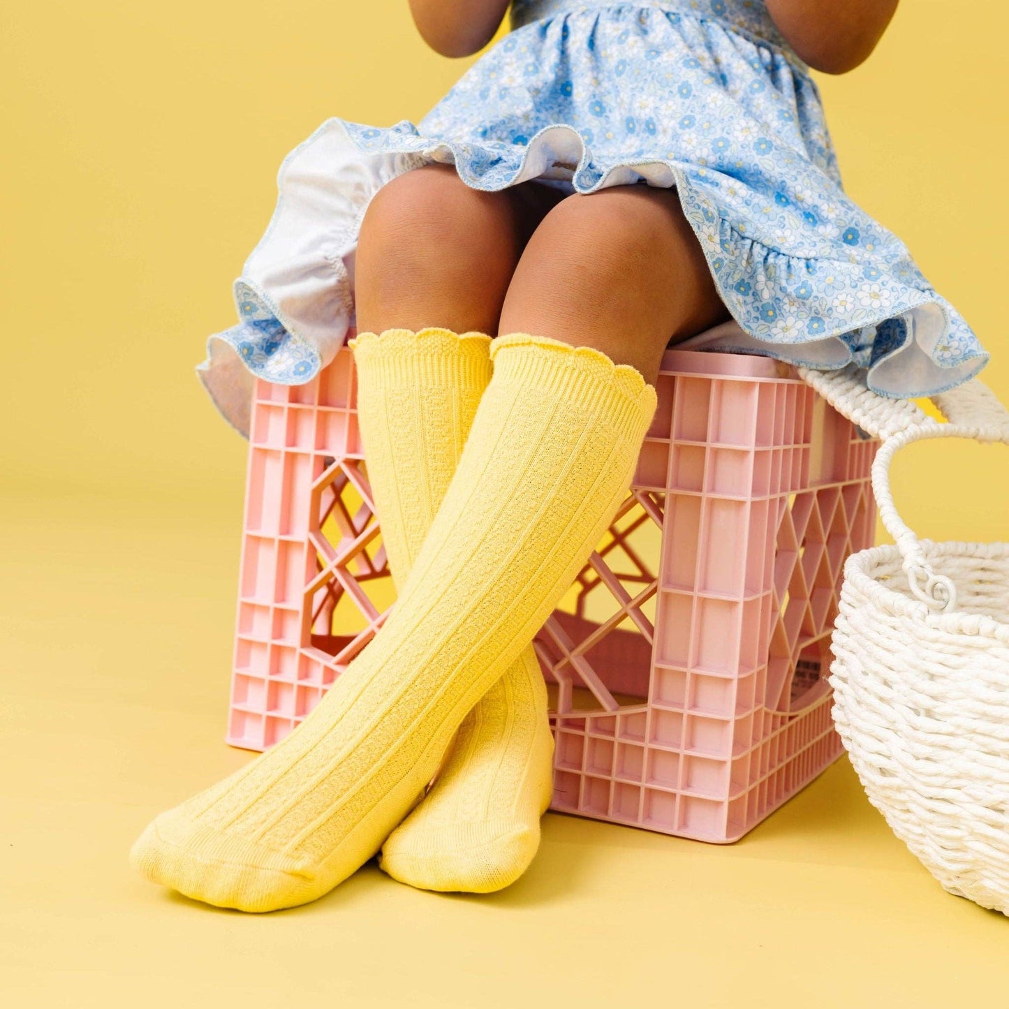 Child wearing yellow knee-high socks and a blue floral dress sitting on pink crates against a yellow background.