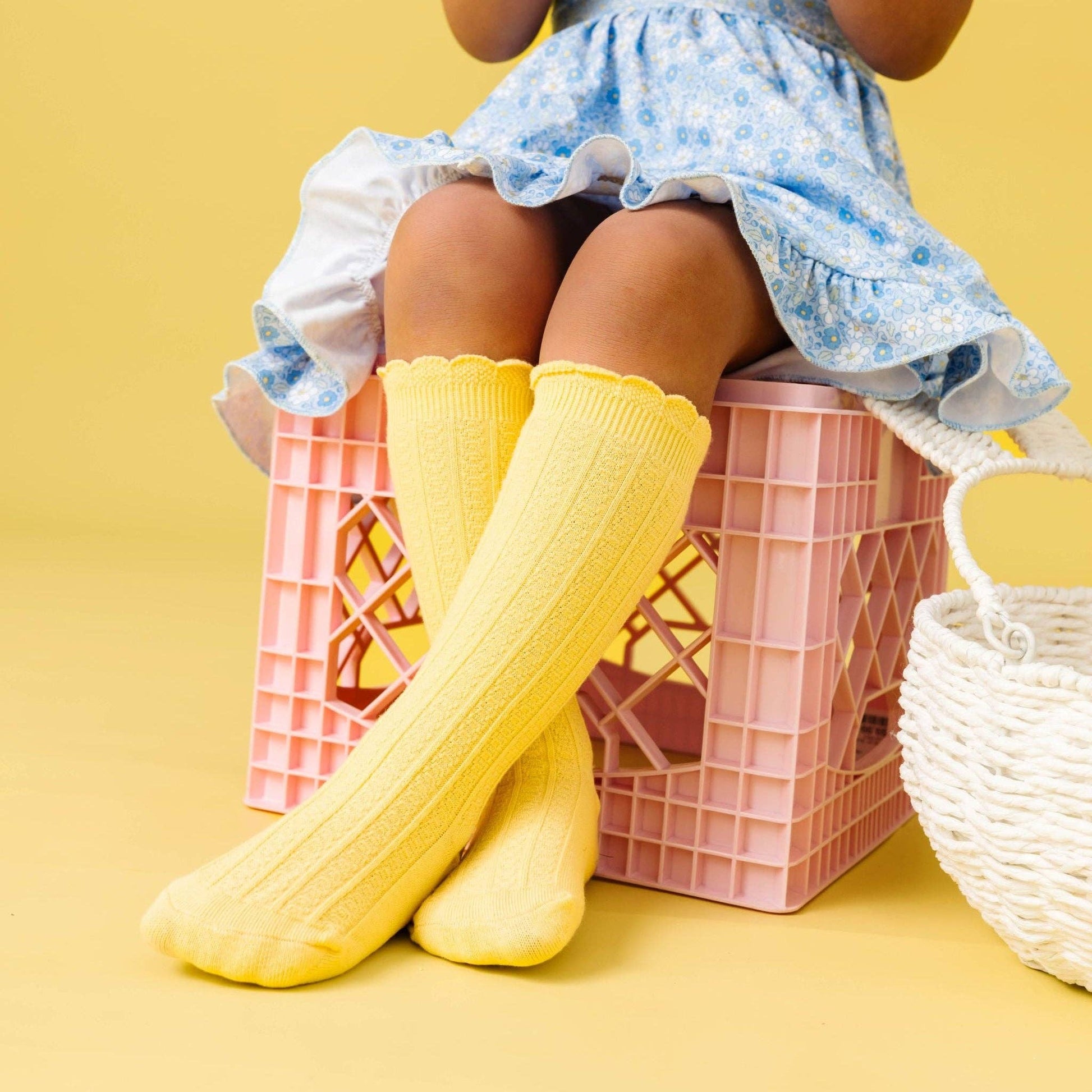 Child wearing yellow knee-high socks and a blue floral dress sitting on pink crates against a yellow background.