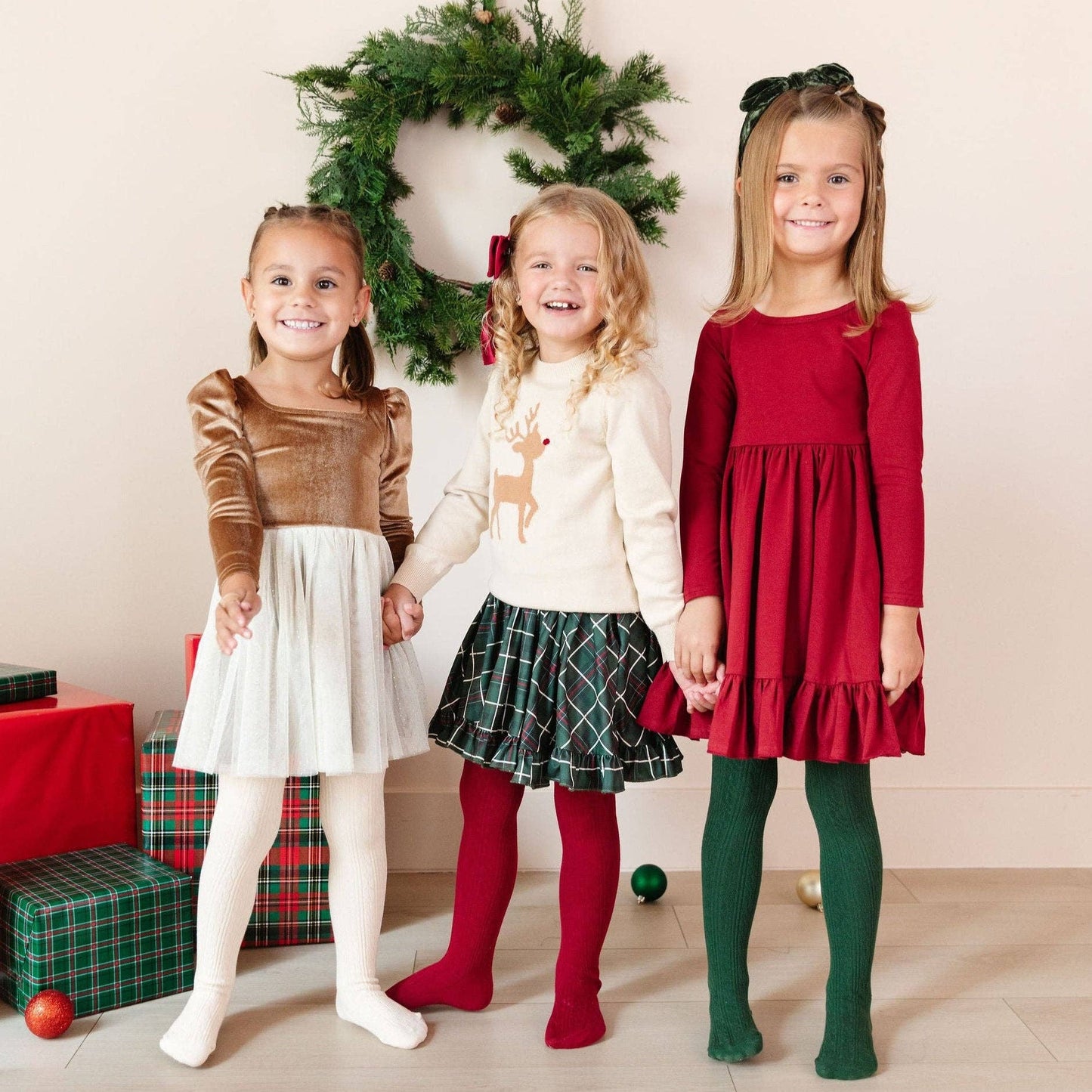 Three young girls in festive outfits standing in front of a Christmas wreath.