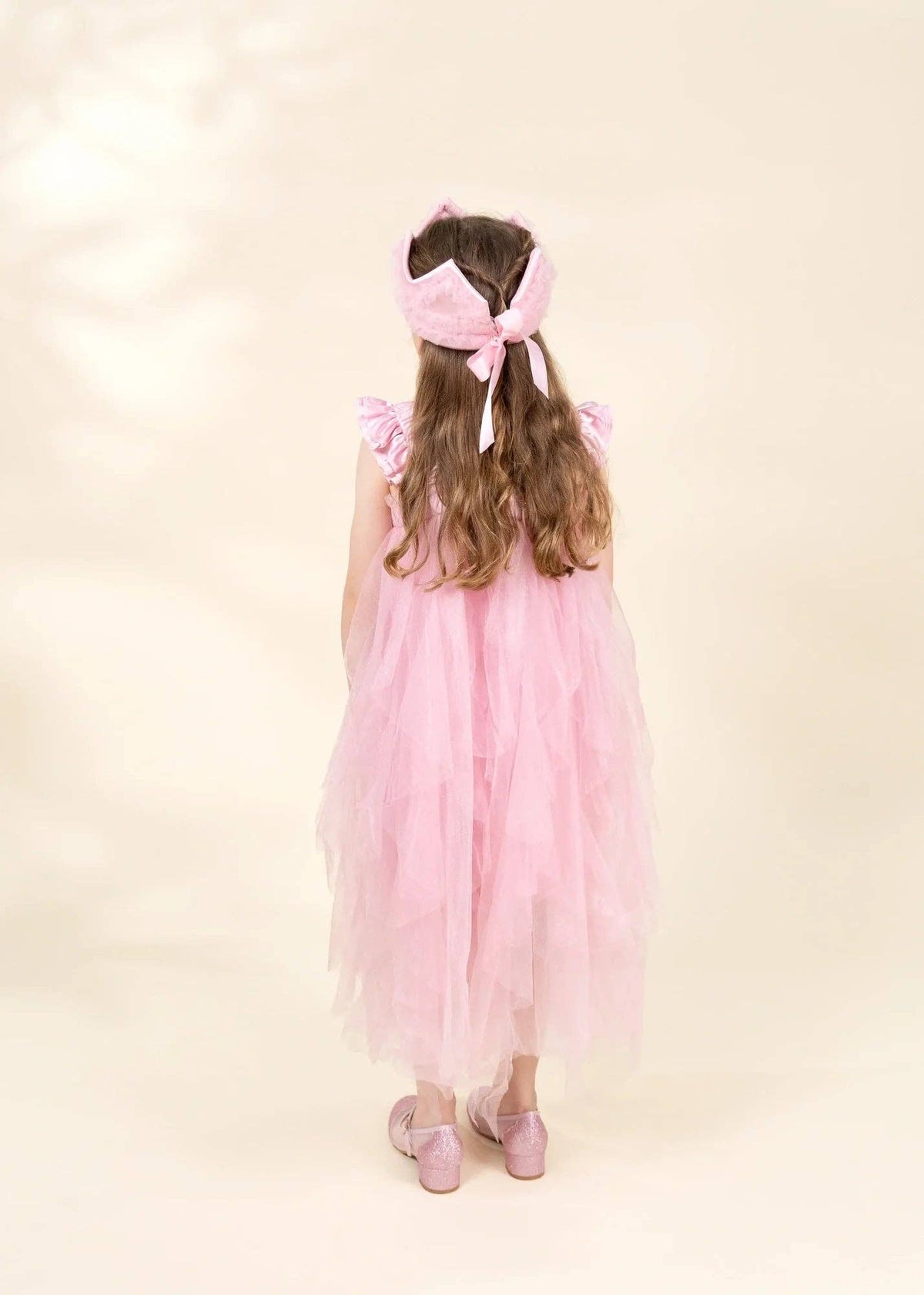 A young girl viewed from behind wearing a pink tiered tulle dress, a matching pink bow headband, and pink ballet slippers, standing against a warm cream background.