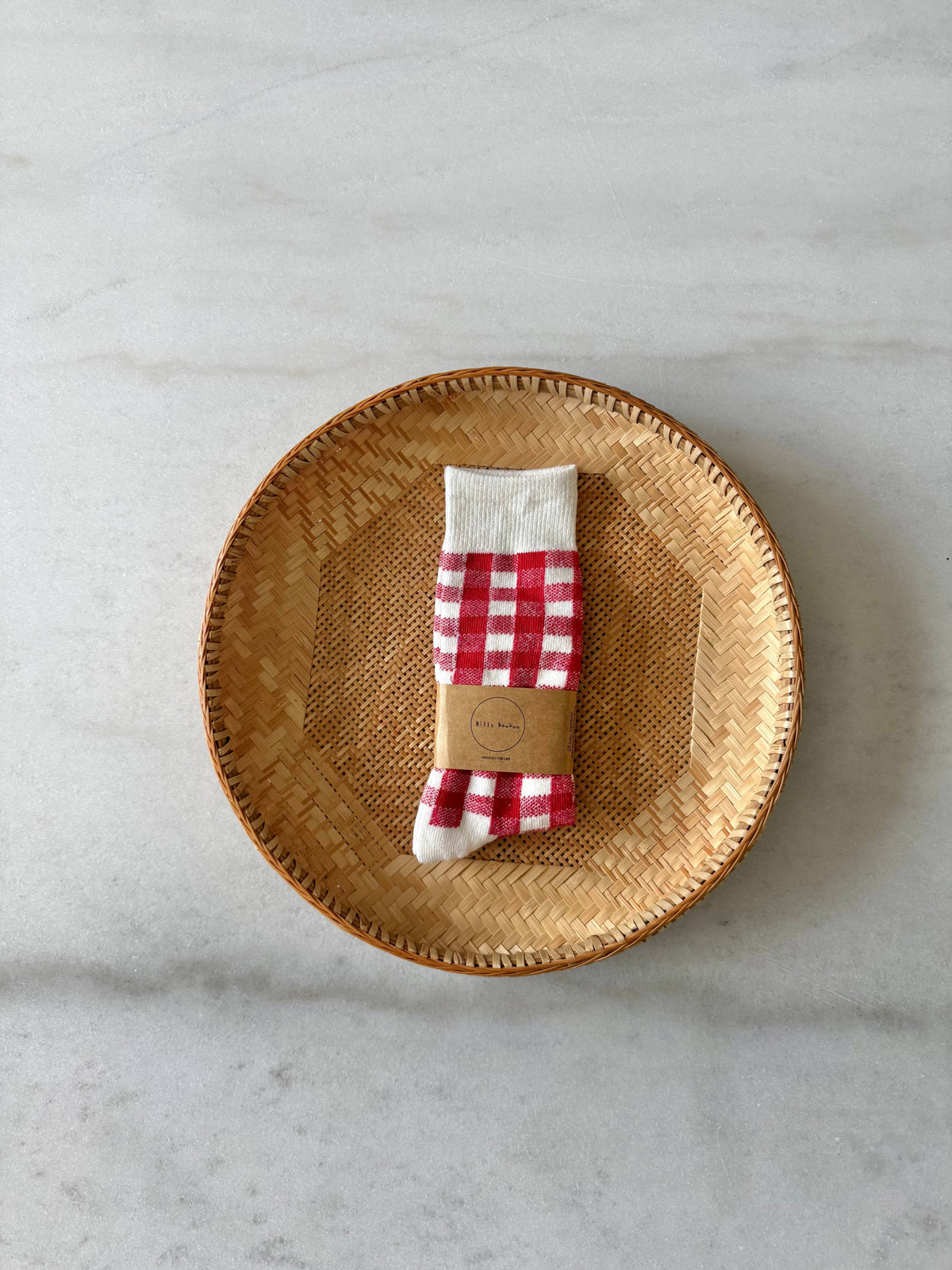 A pair of folded red and white gingham socks with a white cuff, wrapped in a brown paper band, displayed on a round woven bamboo plate over a light gray marble surface.