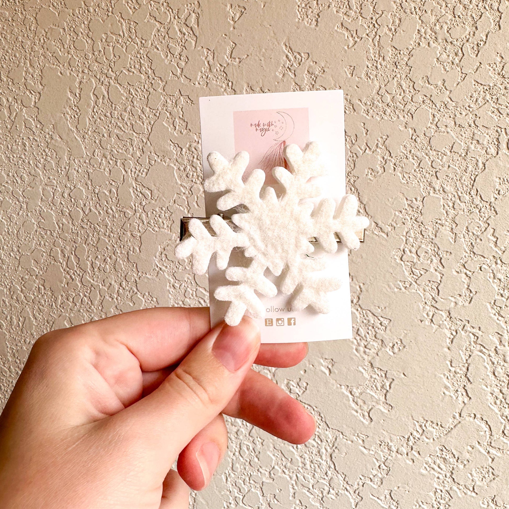 Hand holding a card with snowflake hair clip against a beige wall.
