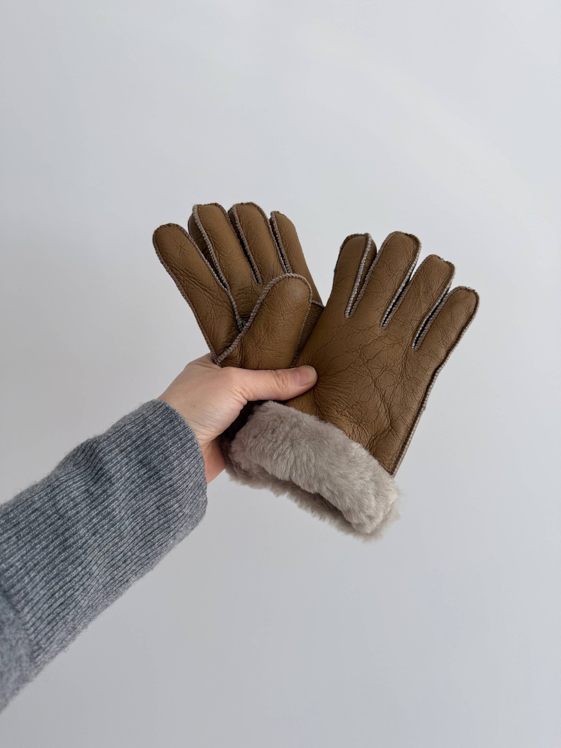 Brown gloves with fur lining held by a person wearing a gray sleeve on a light gray background