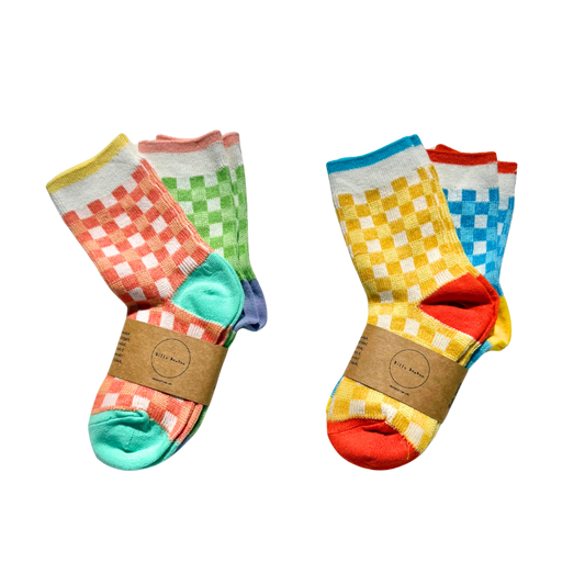 Two pairs of crew socks with colorful checkerboard patterns and solid toe/heel sections, each wrapped with a brown paper band. The left pair features orange, green, and teal colors, while the right pair features yellow, blue, and red colors.