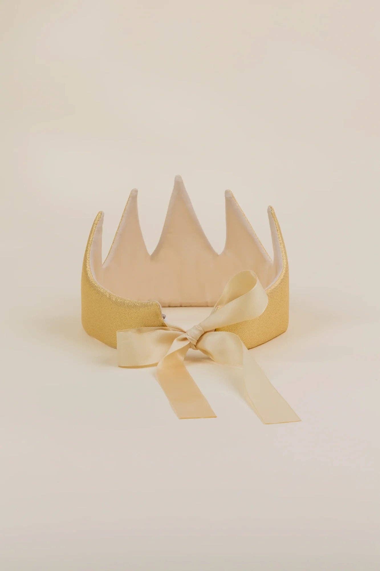 A gold crown-shaped headband with a matching satin ribbon bow, displayed on a neutral cream background.