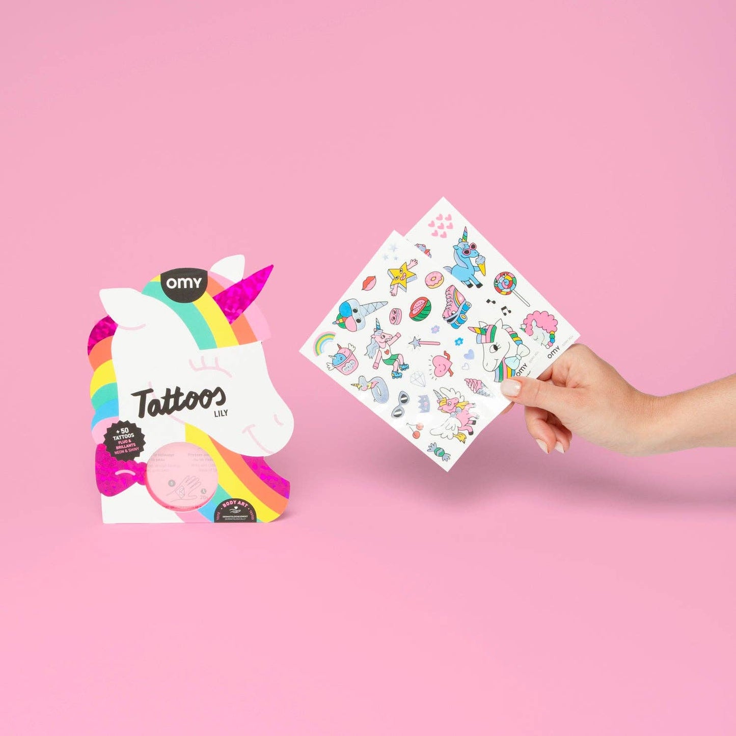 Hand holding a sheet of unicorn-themed tattoos with Omy branding on a pink background