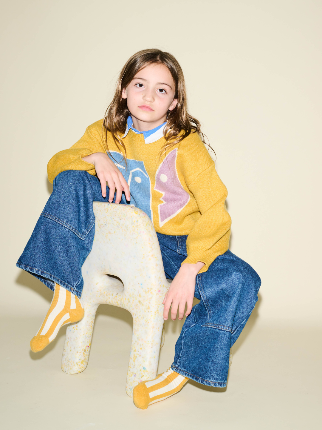 Child wearing a yellow sweater with a colorful design, blue jeans, and yellow socks sitting on a white stool against a beige background.