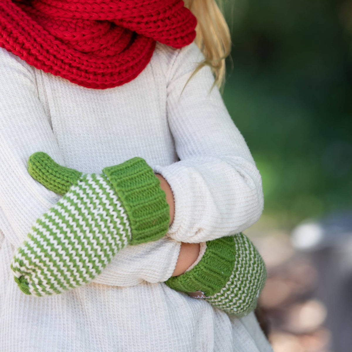 A close-up of a child wearing a thick red knit scarf and white knit sweater, with green and white striped knit mittens on the hands. The background is a soft, blurred outdoor green setting.