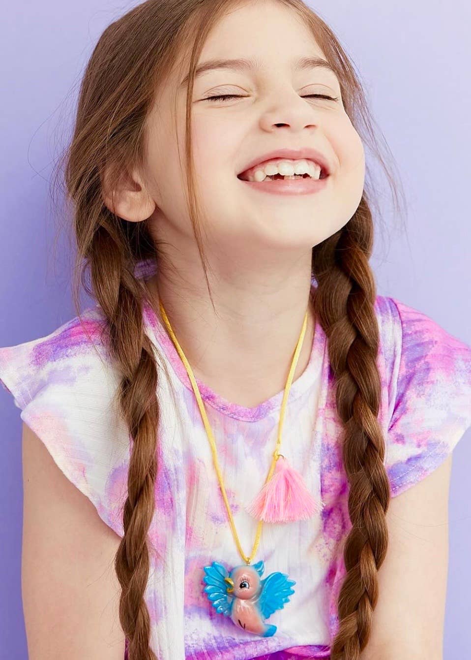 A young girl with long brown braids is smiling with her eyes closed, wearing a colorful tie-dye t-shirt and a yellow cord necklace with a pink and blue bird pendant, set against a solid lavender background.