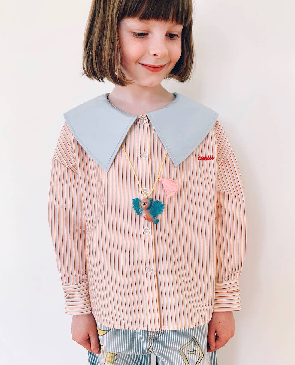 Child wearing a striped shirt with a large collar and colorful necklace on a plain background