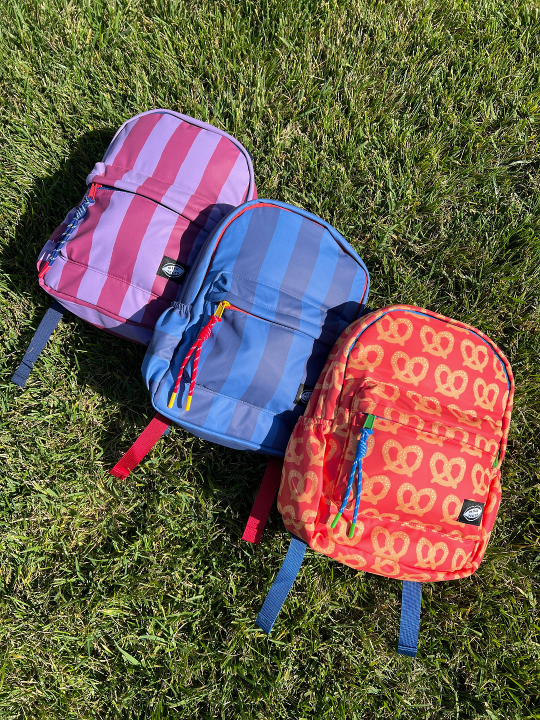 Three small backpacks arranged on green grass in sunlight: one pink and purple striped, one blue and purple striped, and one red-orange patterned with yellow pretzel shapes. Each backpack has blue straps and small black brand patches.