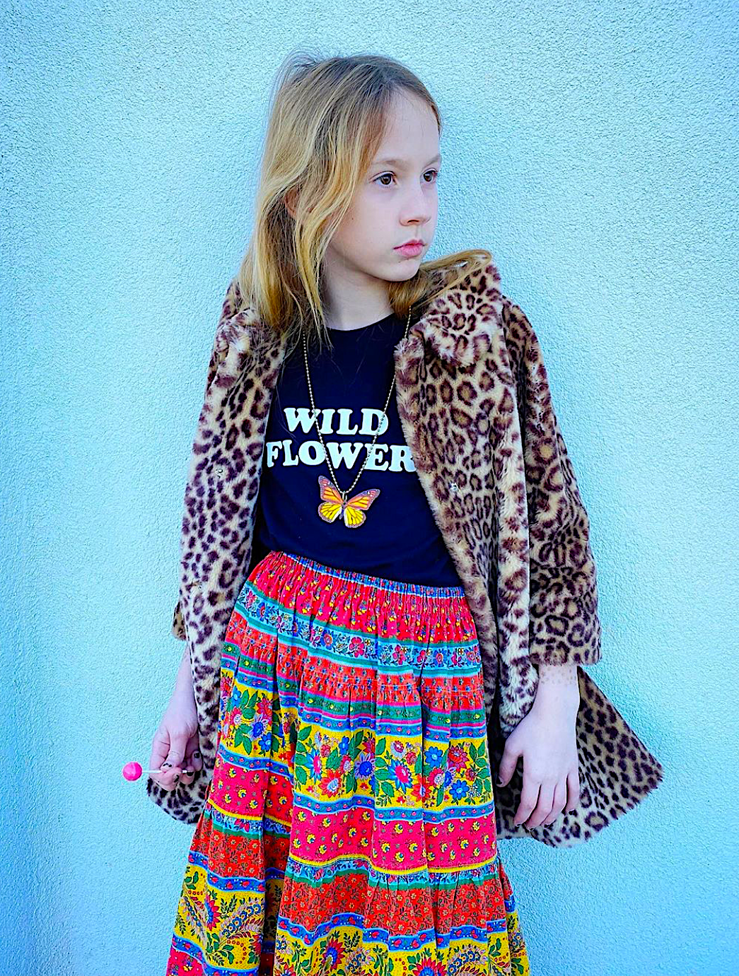 Young girl wearing a leopard print coat, black t-shirt with 'WILD FLOWER' text, and colorful patterned skirt against a light blue background.
