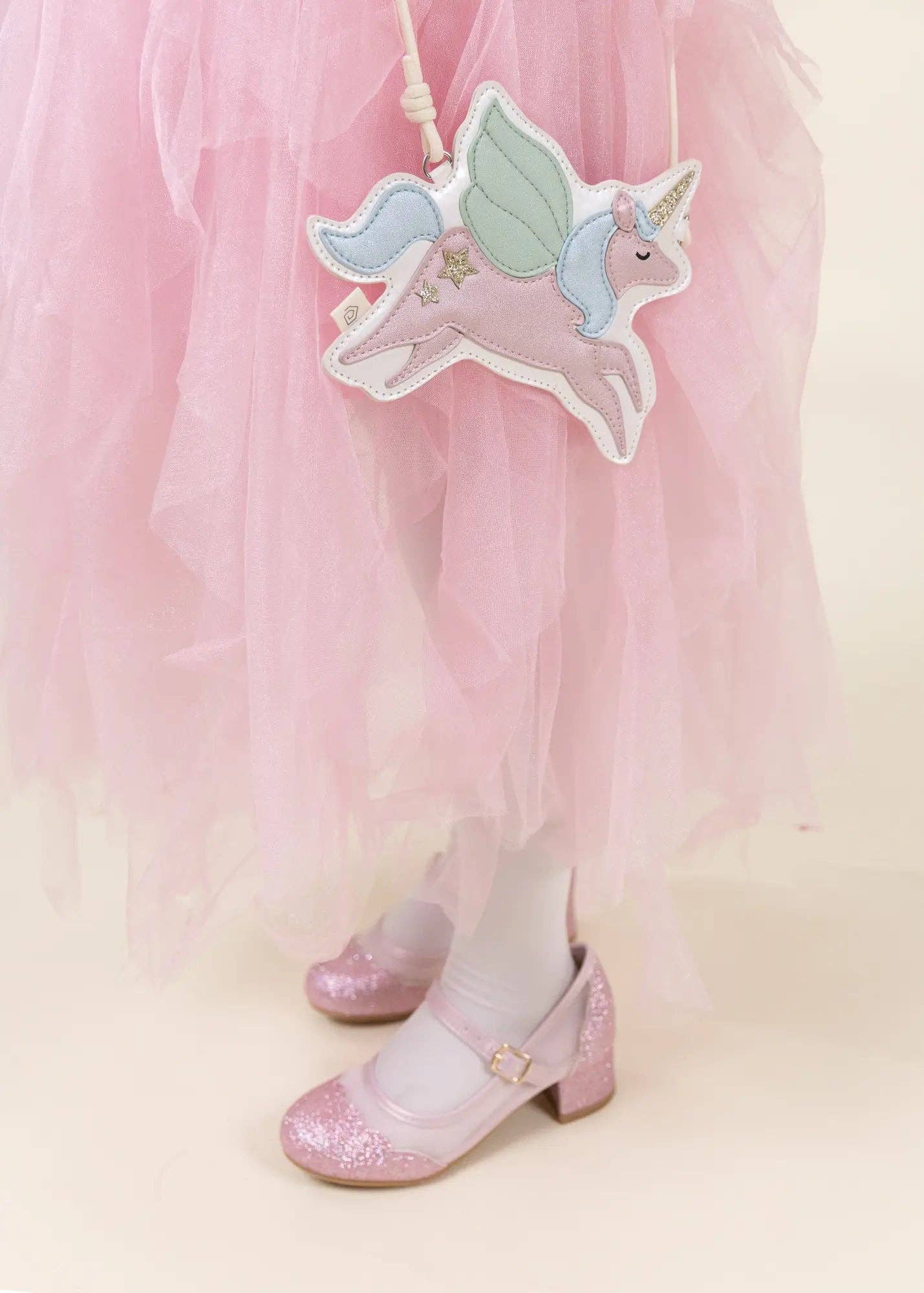 A close-up lifestyle shot of a child's lower legs wearing a fluffy pink tulle tutu skirt, white tights, and sparkly pink Mary Jane shoes, holding a pink unicorn patch bag.