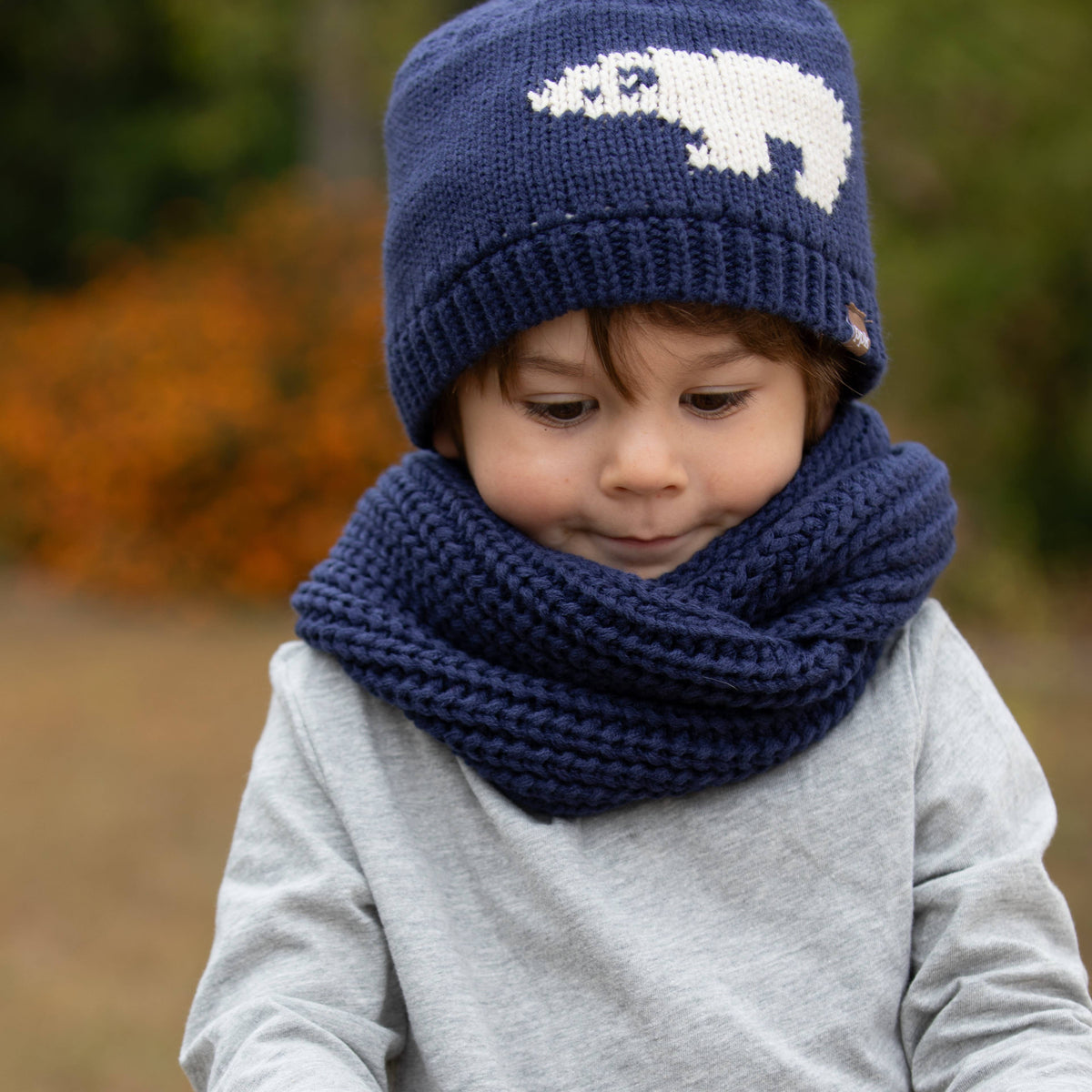 A child wearing a navy blue knit beanie with a white polar bear design and a matching chunky navy cowl neck warmer, photographed outdoors with a blurred natural background.