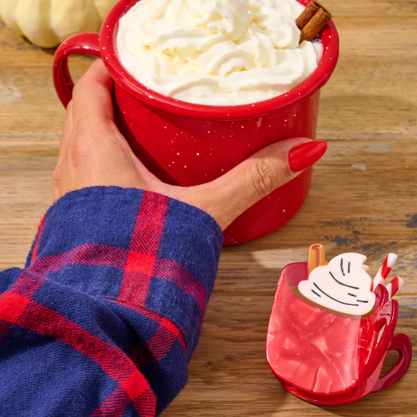 Red mug with whipped cream and a cinnamon stick, held by a person in a blue and red plaid shirt, on a wooden surface.