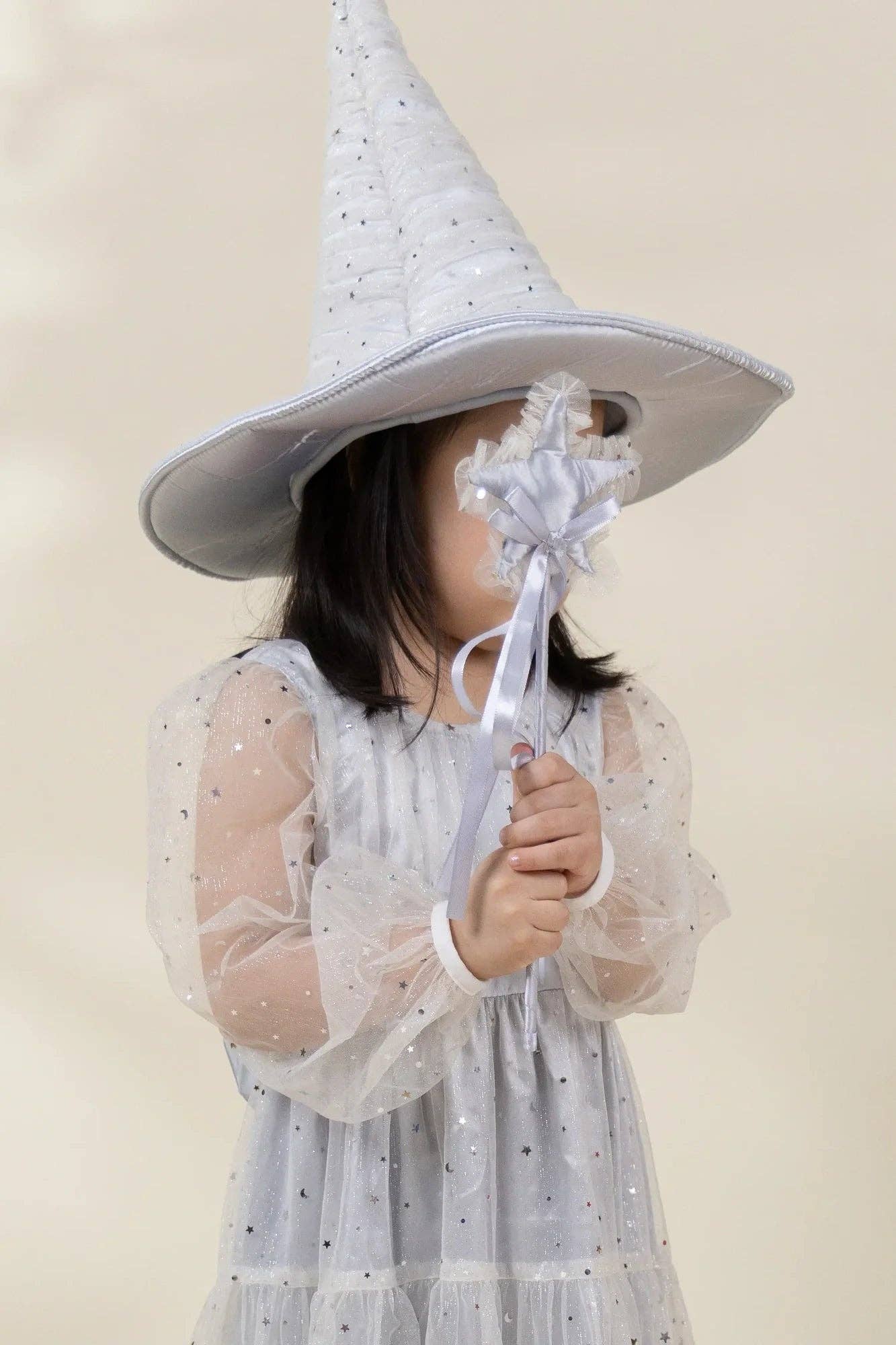 A child wearing a tall pointed white witch hat with small silver star details and a matching white tulle dress with star accents, holding a white wand with a flower to block part of the face, against a soft beige background.