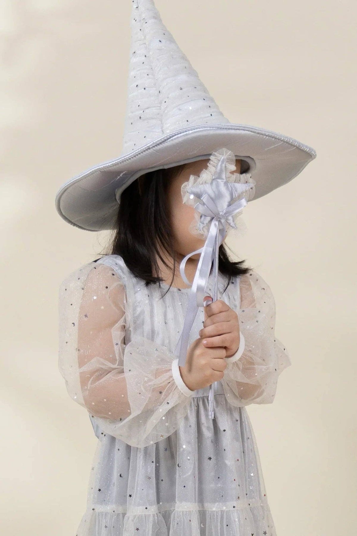 A child wearing a tall pointed white witch hat with small silver star details and a matching white tulle dress with star accents, holding a white wand with a flower to block part of the face, against a soft beige background.