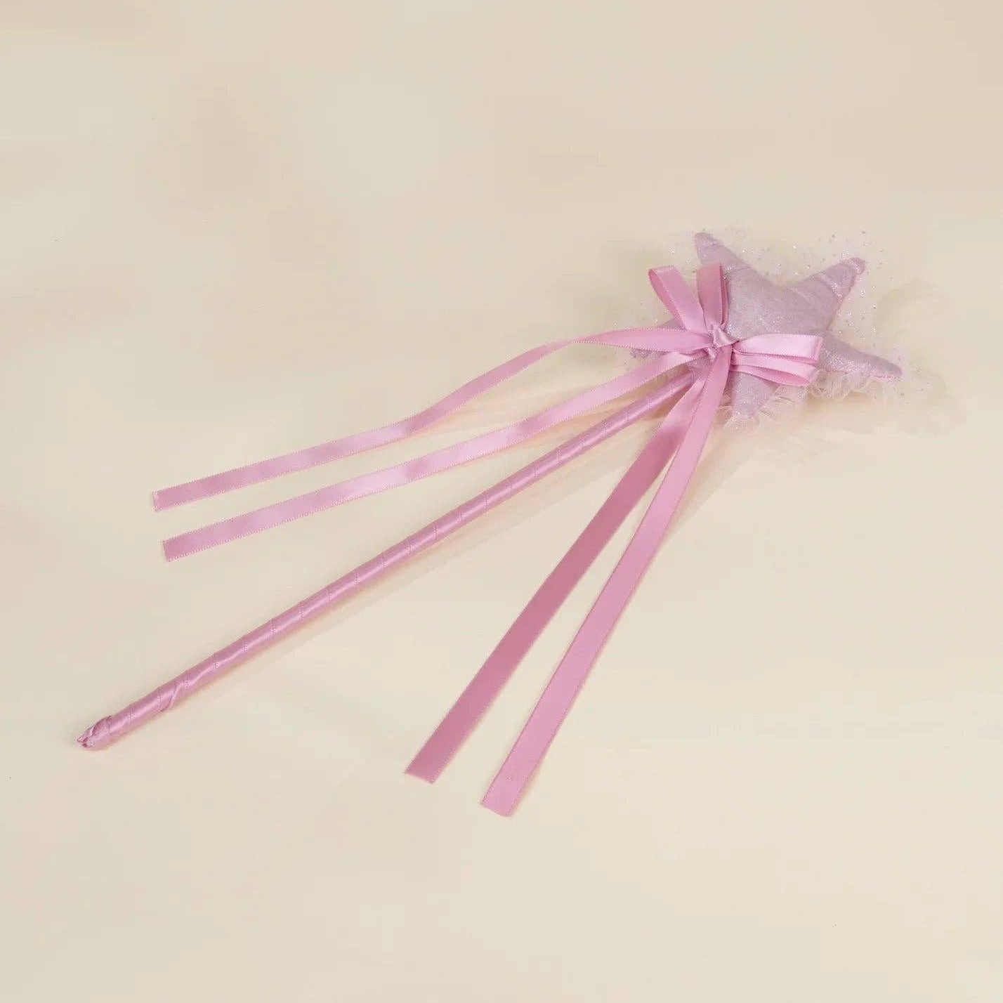 A pink star-shaped wand with long satin ribbons and a small tulle bow, photographed on a plain light beige background.