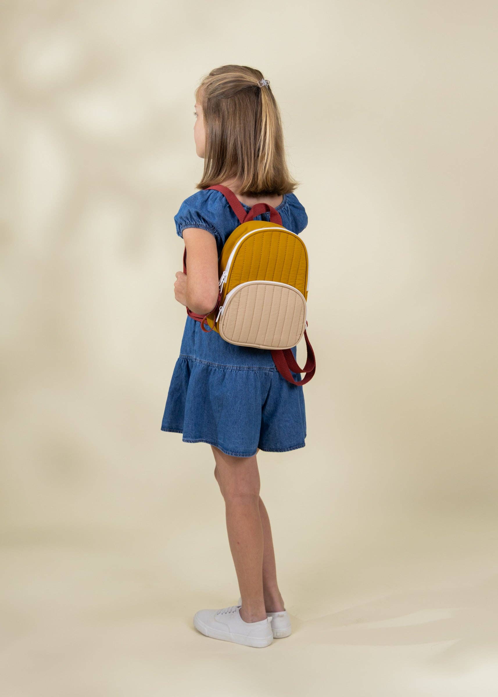 Young girl wearing a blue dress and yellow backpack on a beige background