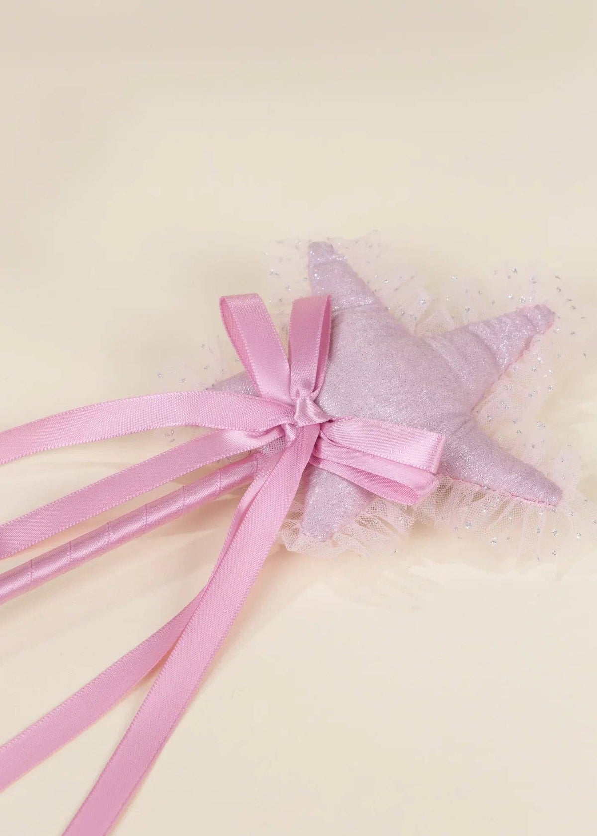 A pink fabric star wand topper with a glittery lavender star center, a pink satin bow, and long pink ribbon tails, shown on a light cream background.
