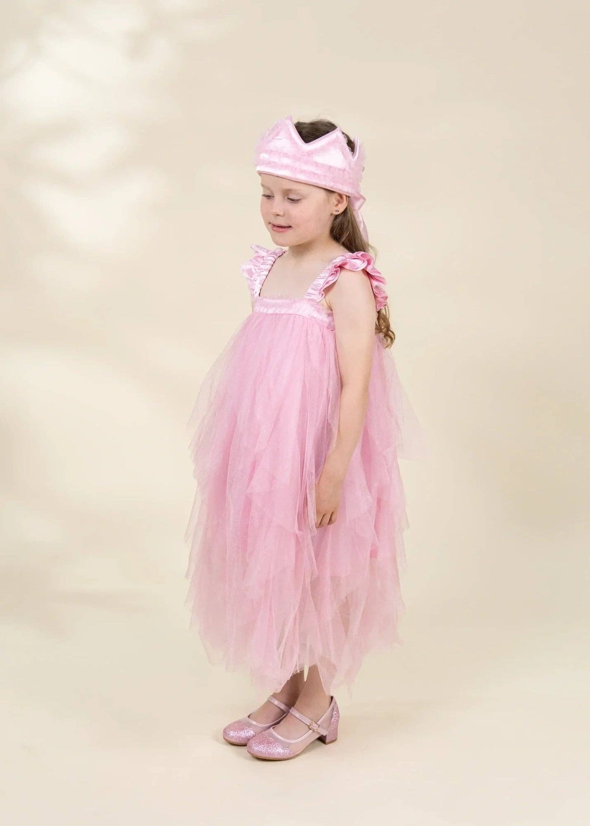 A young girl wearing a light pink tulle dress with ruffled straps, a matching pink headband, and pink shoes, standing against a plain beige studio background.