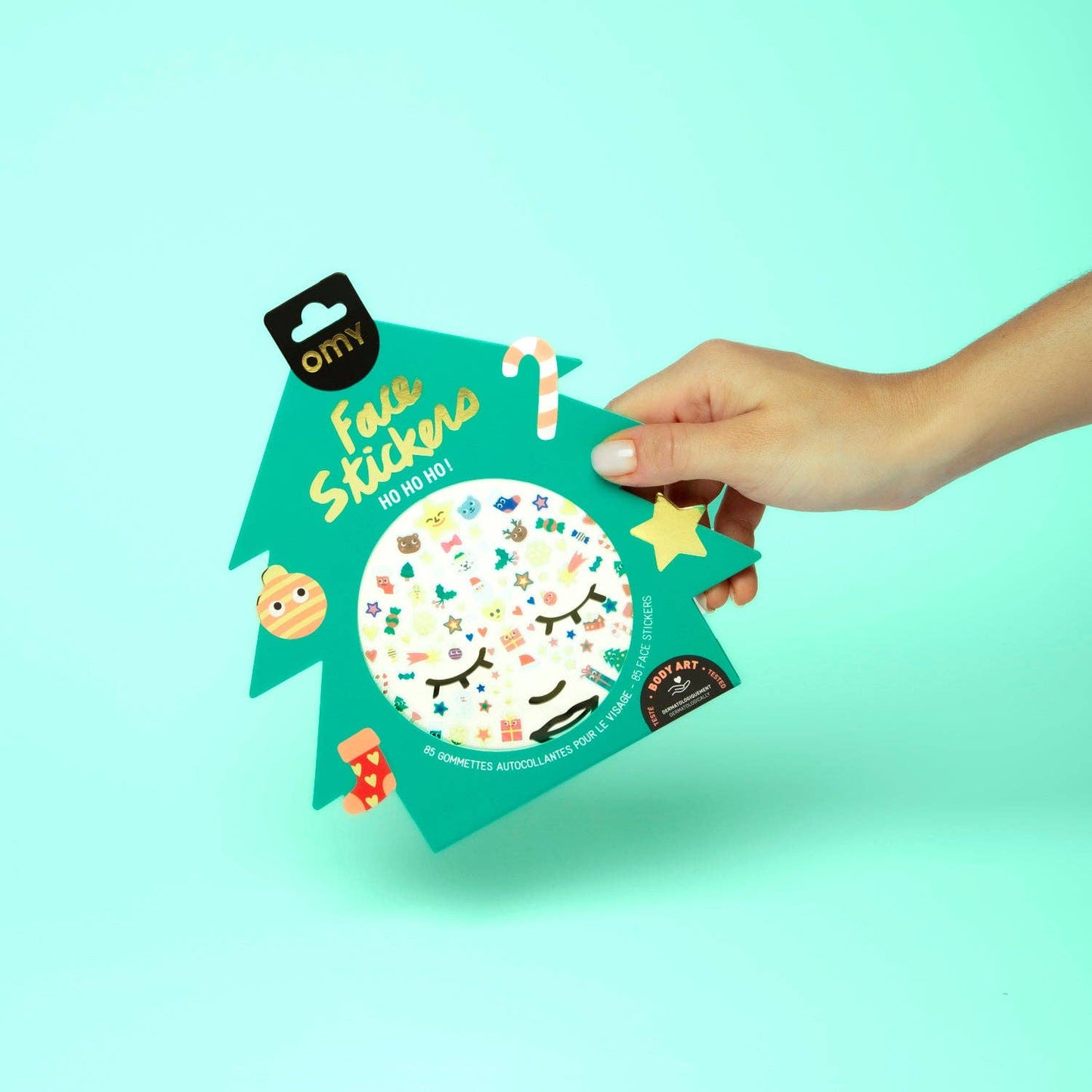 Hand holding a package of Omy face stickers against a light blue background