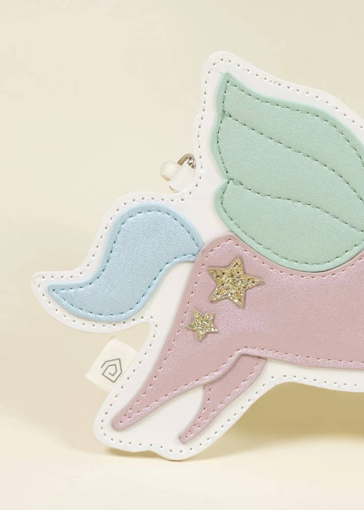 A pastel unicorn-shaped accessory with stitched fabric wings in mint green and light blue, a pink horn, and two small gold glitter stars, shown on a warm cream background.