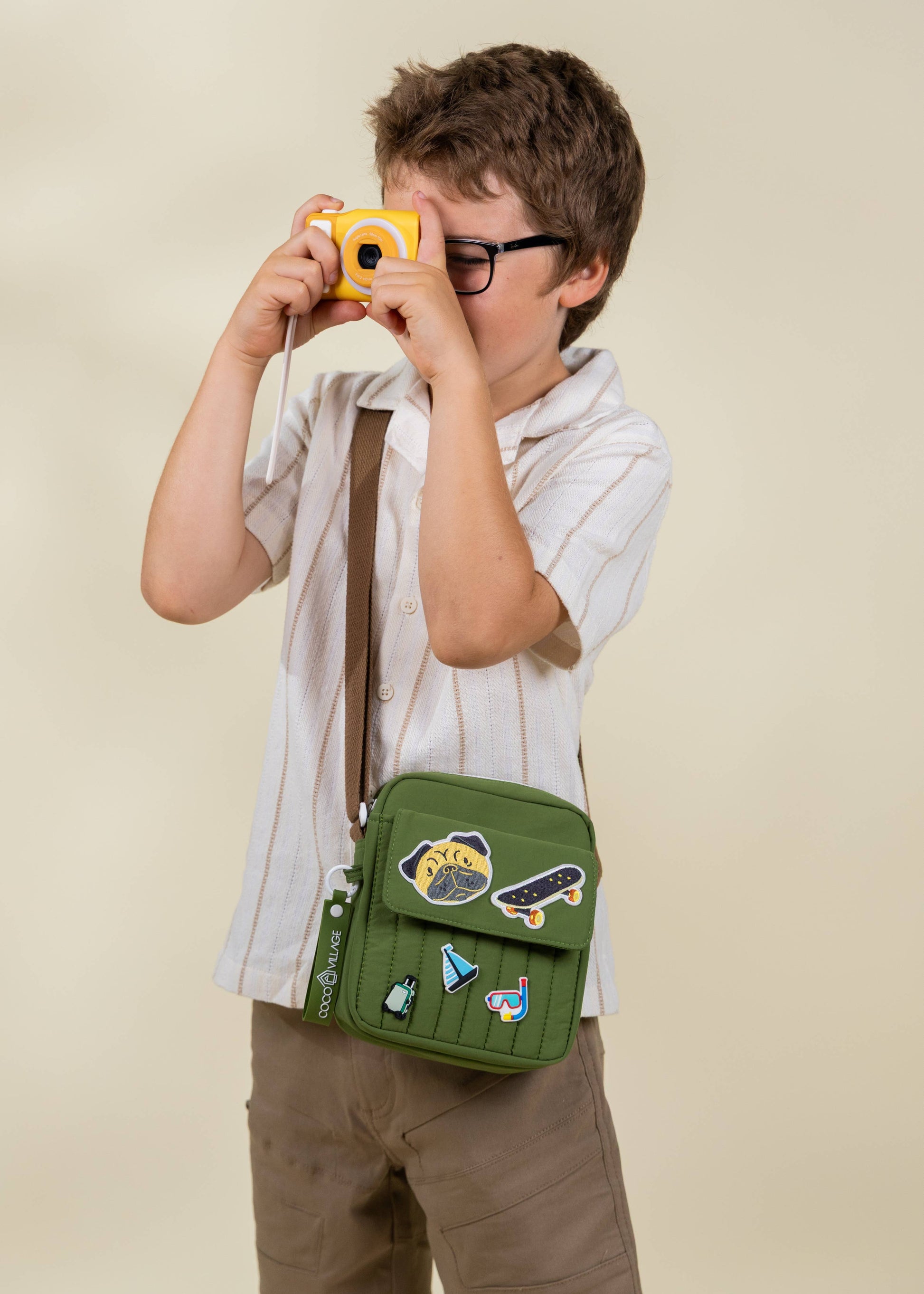 Child holding a green bag with patches and taking a photo with a yellow camera.
