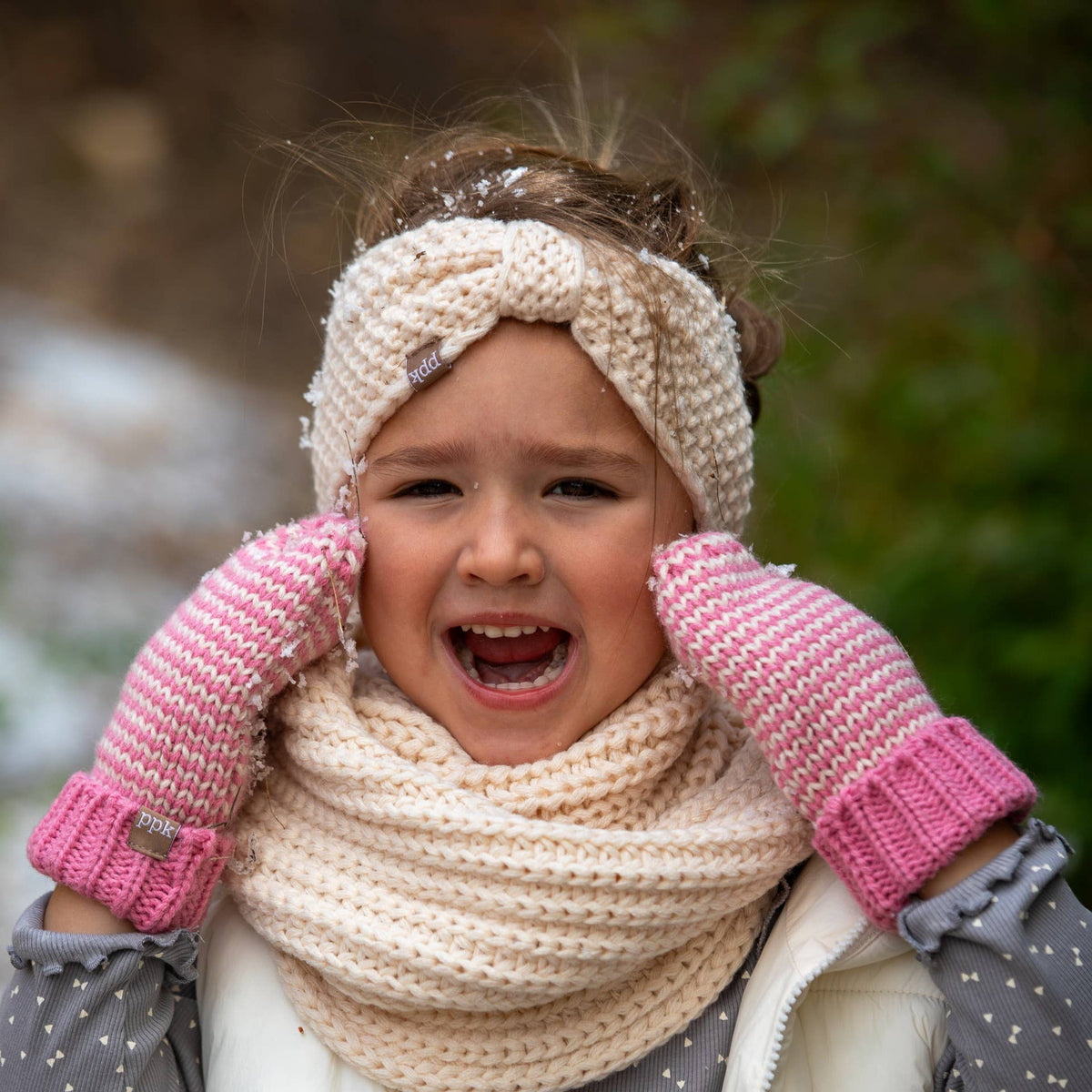 A young child wearing a cream knit headband, matching knit scarf, and pink knit mittens, photographed outdoors with a blurred natural background.