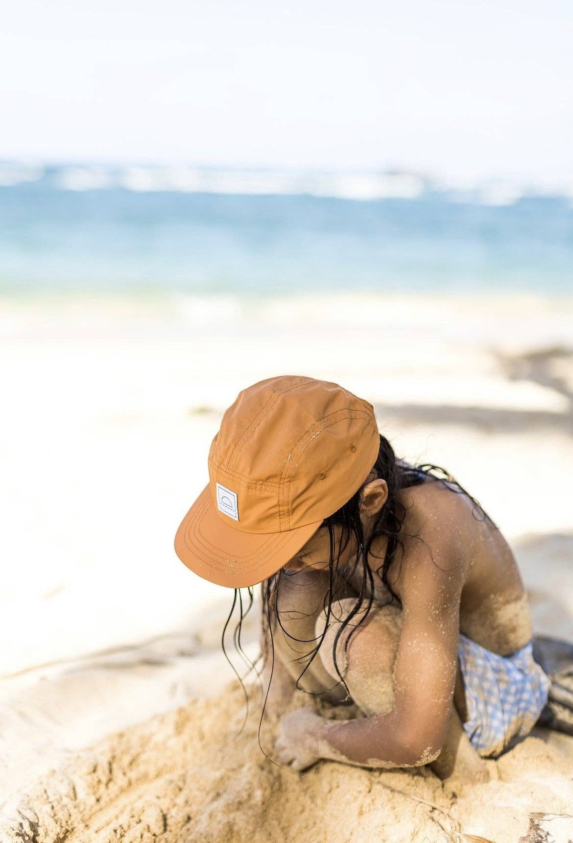 Person wearing a brown cap sitting on sand with a blurred beach background