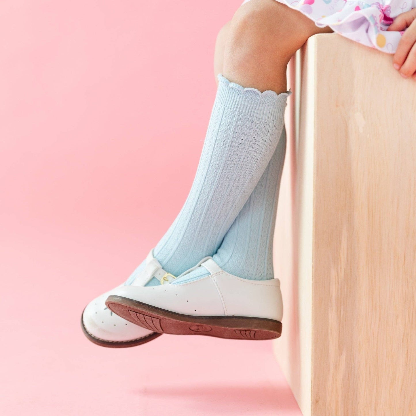 Child's legs wearing light blue socks and white shoes on a pink background