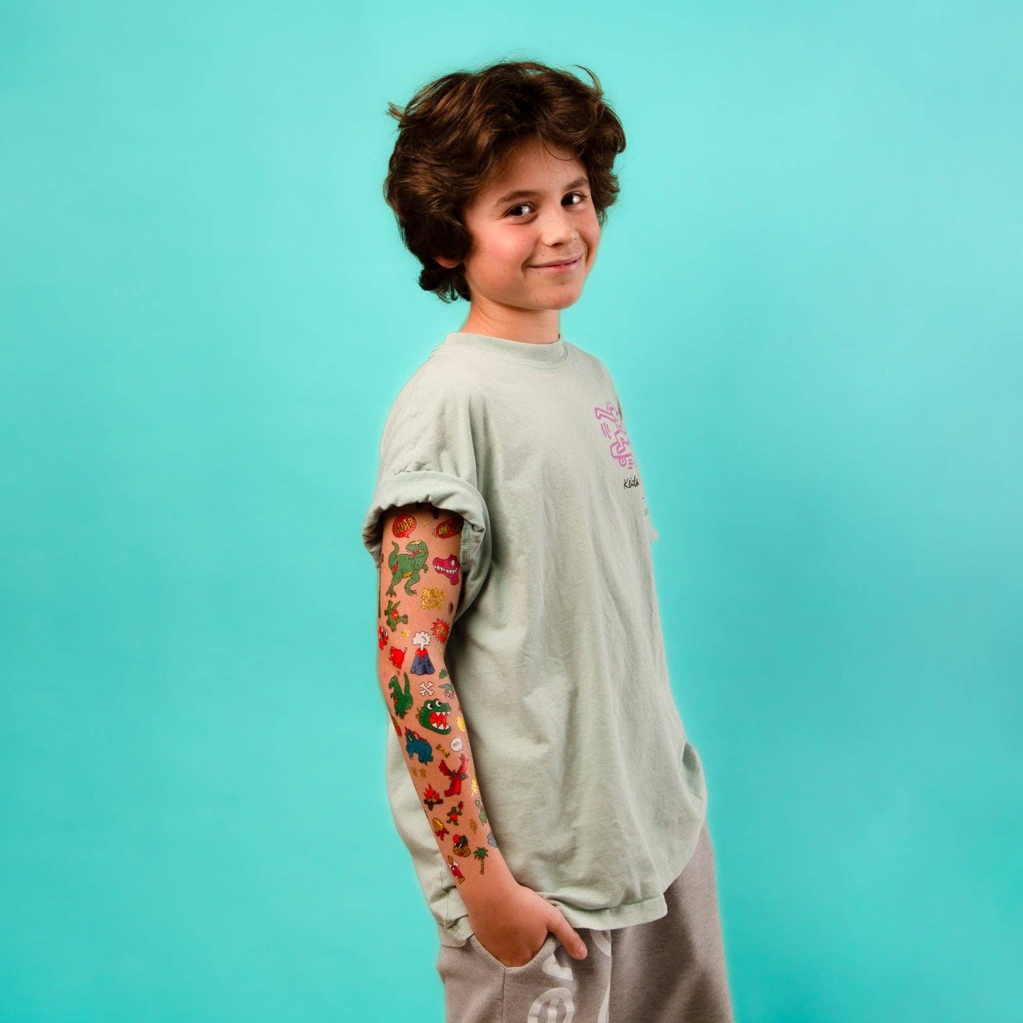 Child with tattoos on arm wearing a light gray t-shirt against a teal background