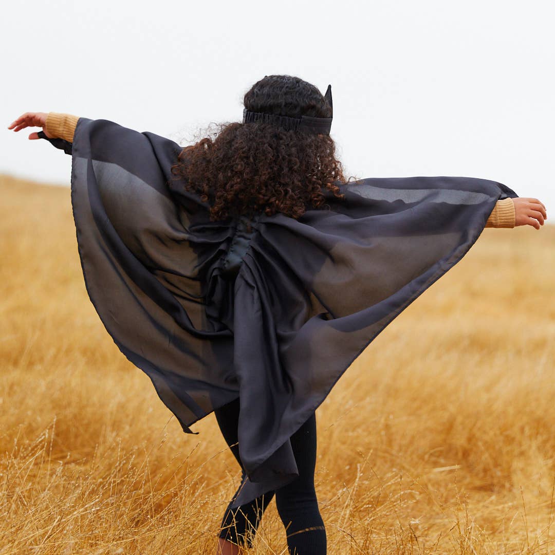 A woman stands in a golden field with her back to the camera, wearing a flowing sheer black cape or poncho with attached bat-like headpiece and black leggings.