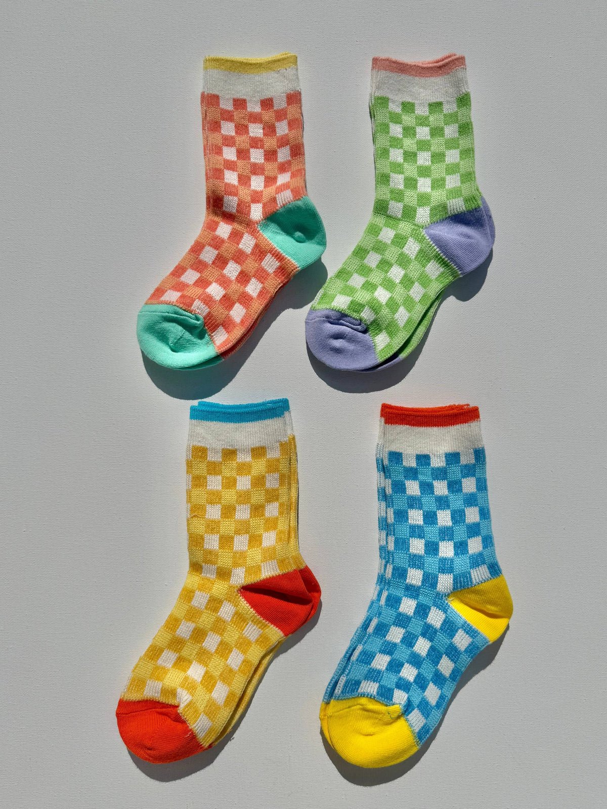 Four crew socks arranged in a 2x2 grid on a light gray background, each featuring a checkered pattern in a different color: orange and white, green and white, yellow and white, and blue and white. The toes, heels, and cuffs of the socks are solid colors in teal, purple, red, orange, yellow, and blue respectively.