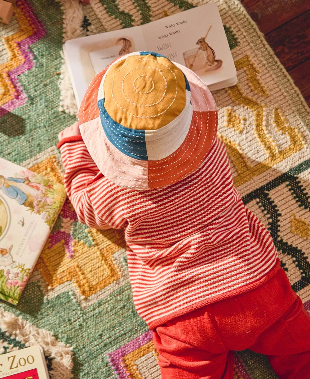 Child wearing a colorful patchwork hat and red outfit on a patterned rug with books around