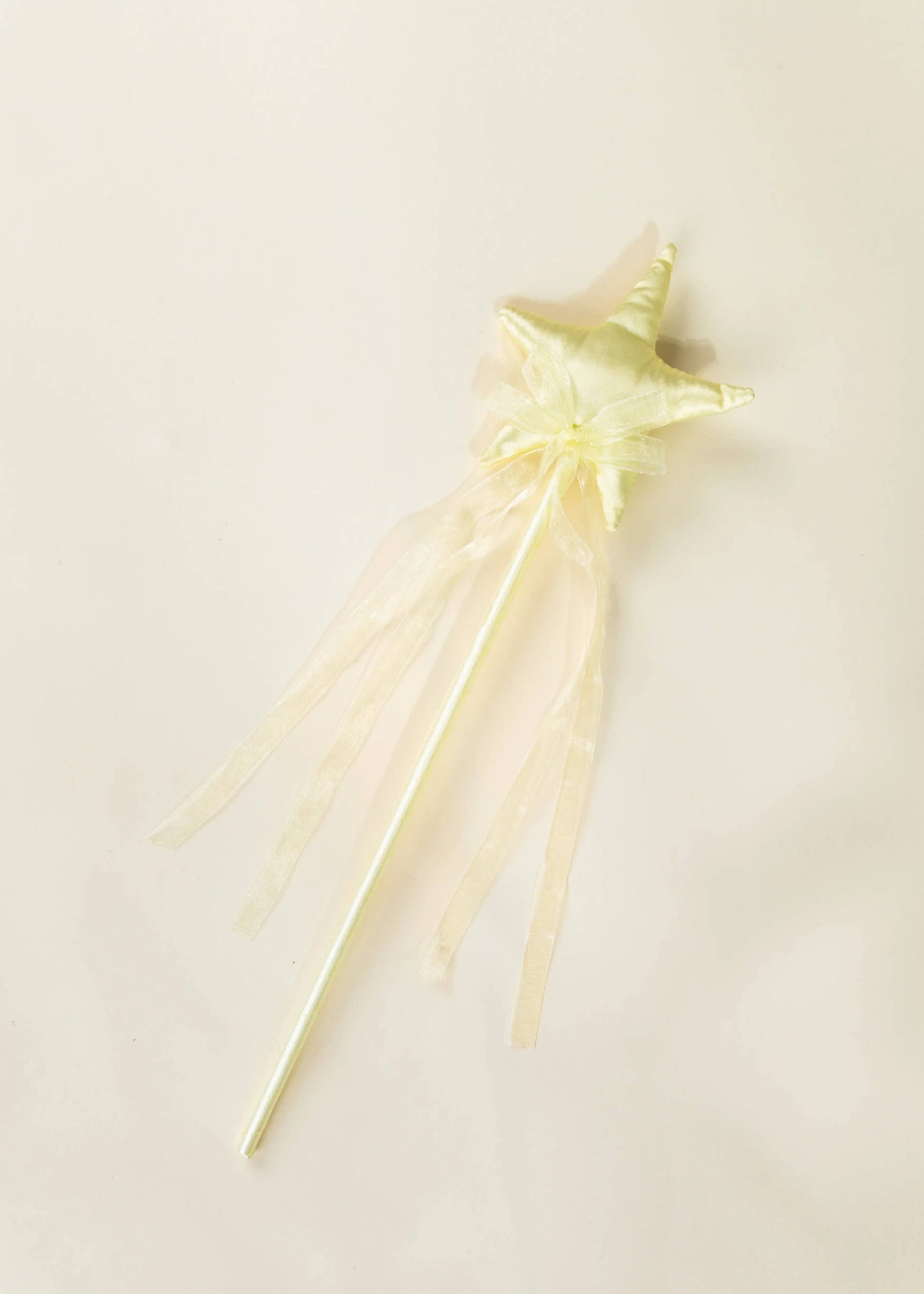 A pale yellow star-shaped toy wand with a matching bow and long sheer ribbon tails, photographed on a plain light beige background.