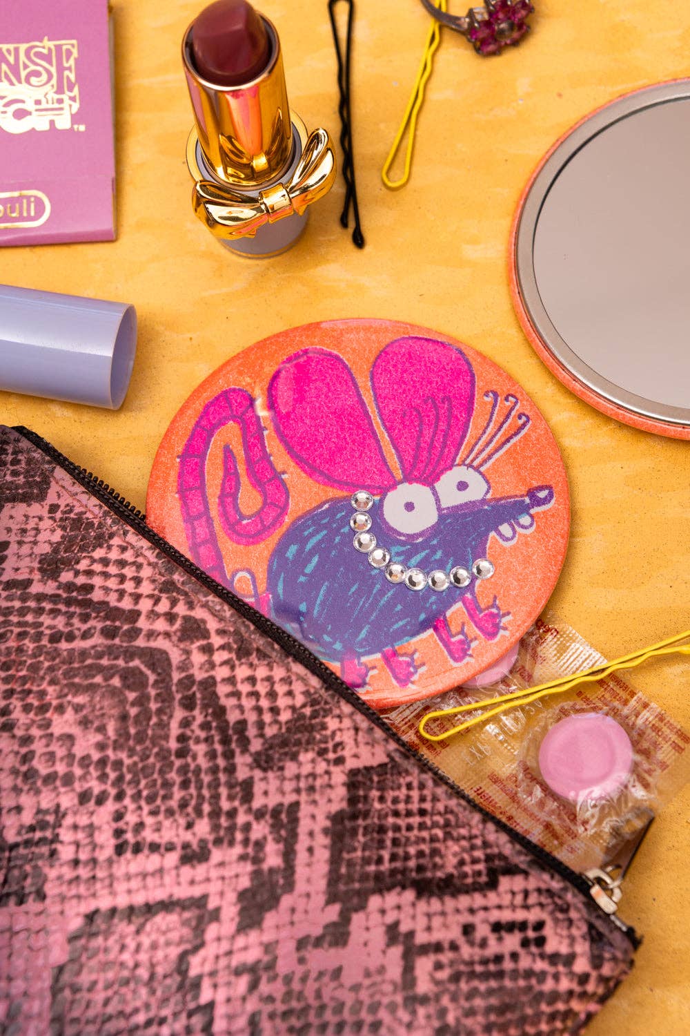 A round orange mouse-shaped makeup sponge with a pink and purple mouse illustration, styled on a yellow surface with various cosmetics including a pink lipstick, a snakeskin pouch, and a compact mirror.