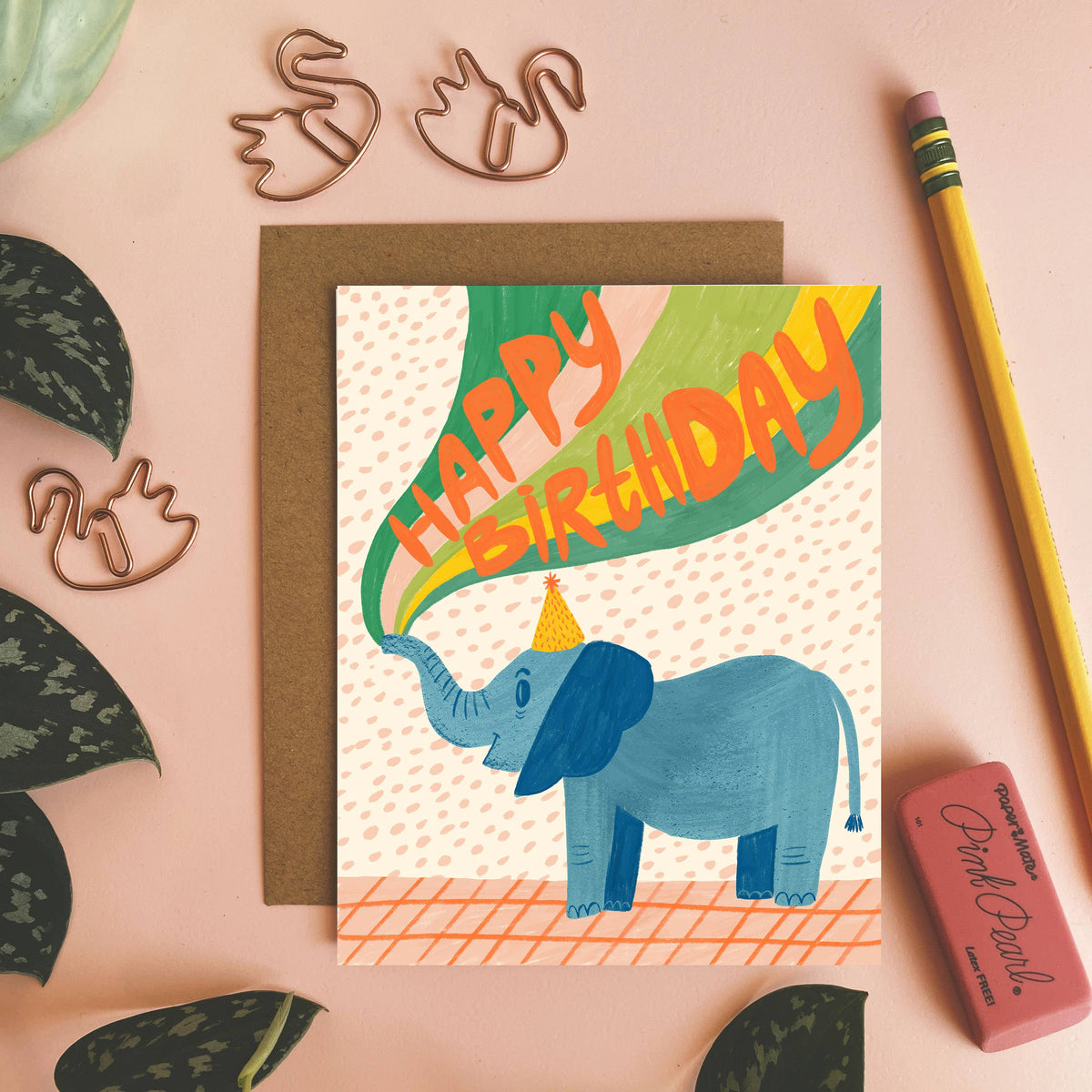 Birthday card with an elephant illustration on a pink surface with decorative elements.