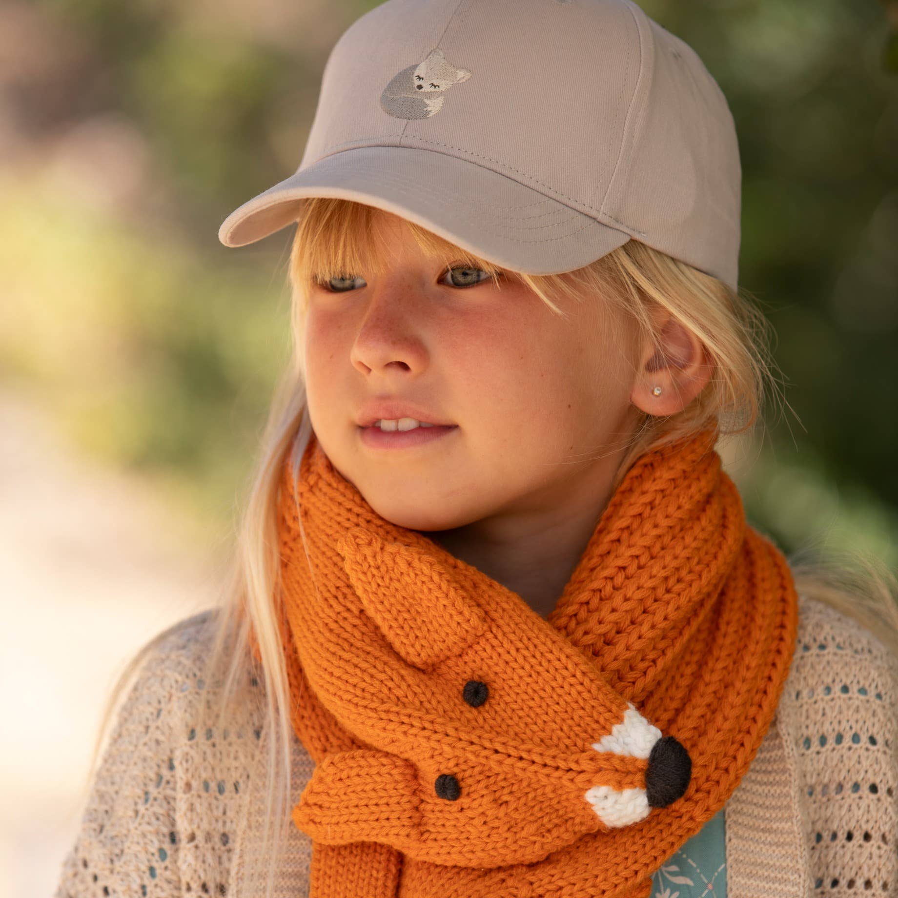 Child wearing a beige cap and orange knitted scarf with a fox design outdoors.