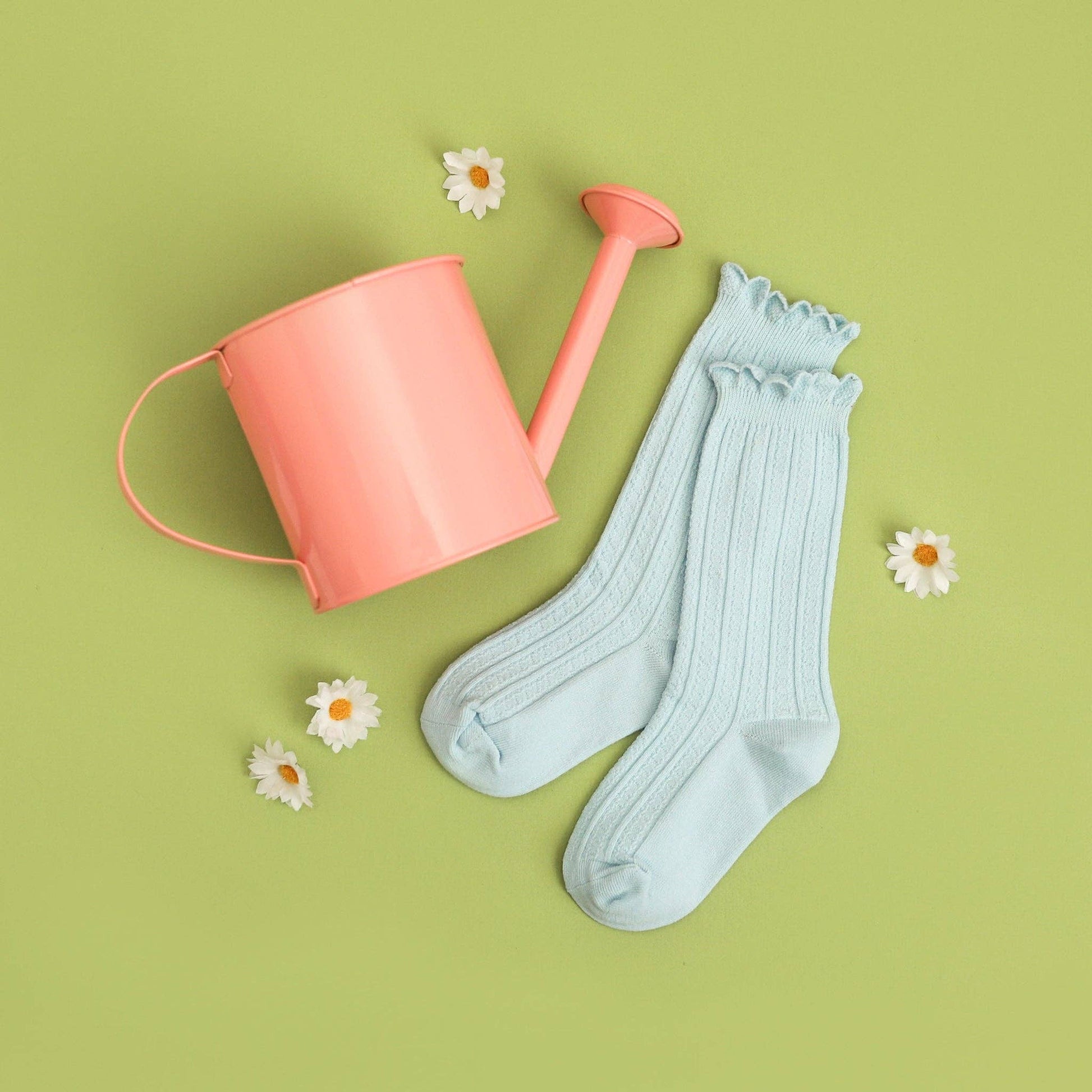 Light blue socks with ruffles next to a pink watering can on a green background