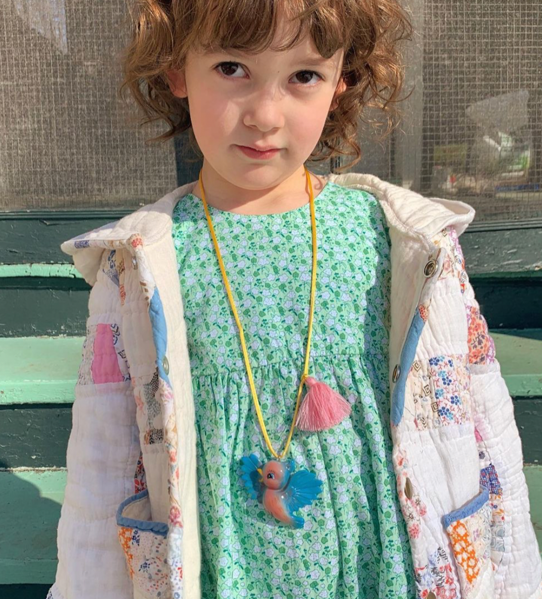 A young child wearing a light green floral dress, a white patchwork quilted jacket, and a yellow cord necklace with a colorful bird charm, standing outdoors in front of a green bench.