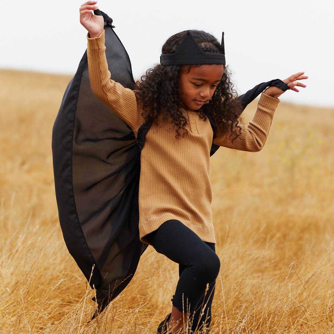 A young girl with curly hair wearing a black bat costume, featuring a headband with horns, a black cape, and a tan ribbed long-sleeve top, posing in a dry grass field.