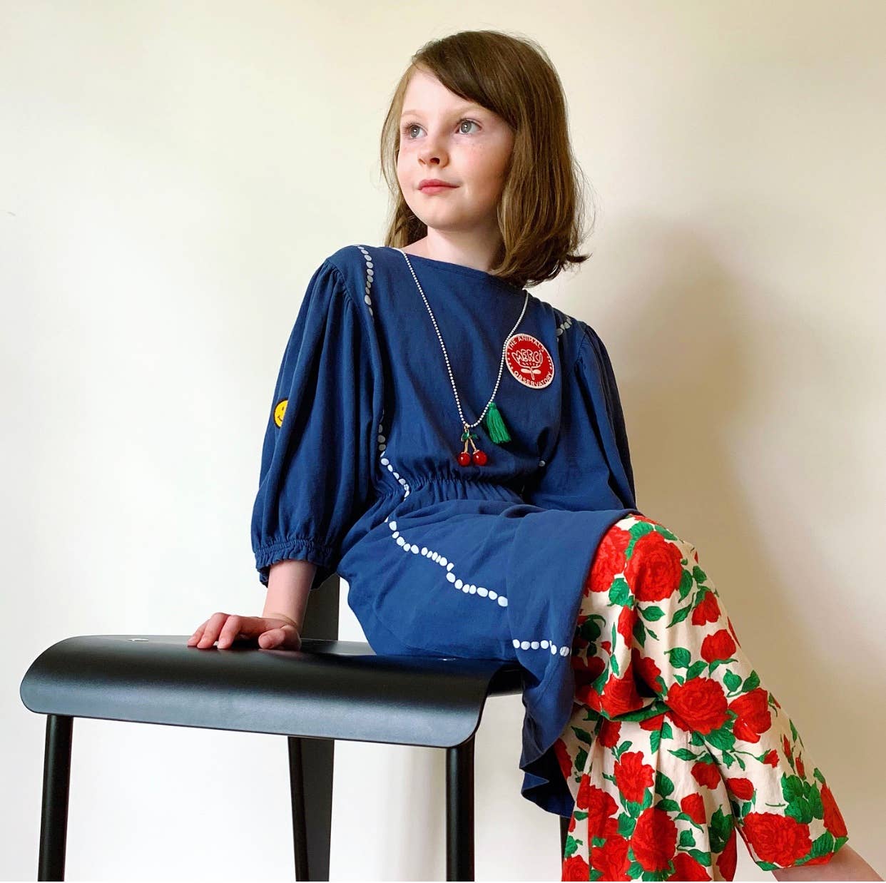 Young girl in a blue dress with a floral shawl, sitting on a black stool against a plain background